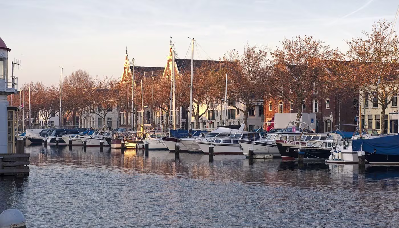 Jachthaven met boten en historische gebouwen aan de waterkant, herfstsfeer met gouden licht.
