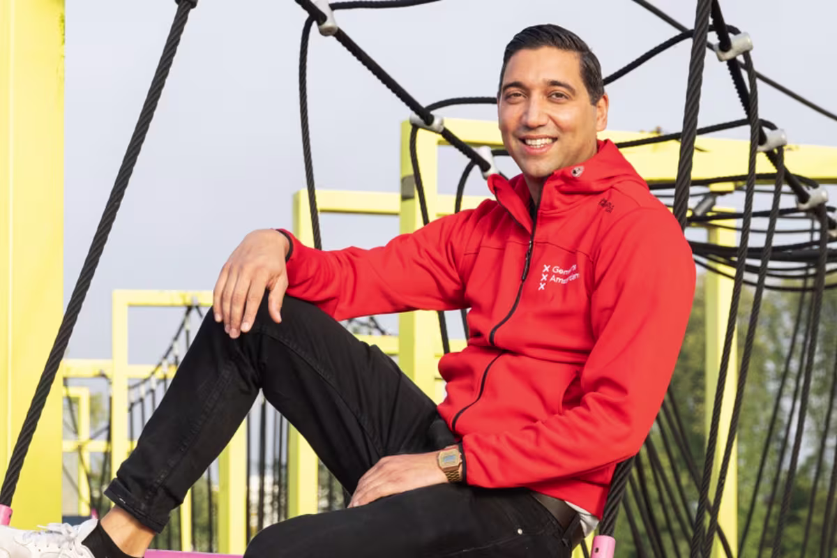 Een man in een rode hoodie zit glimlachend op een geel speeltoestel in een speeltuin.
