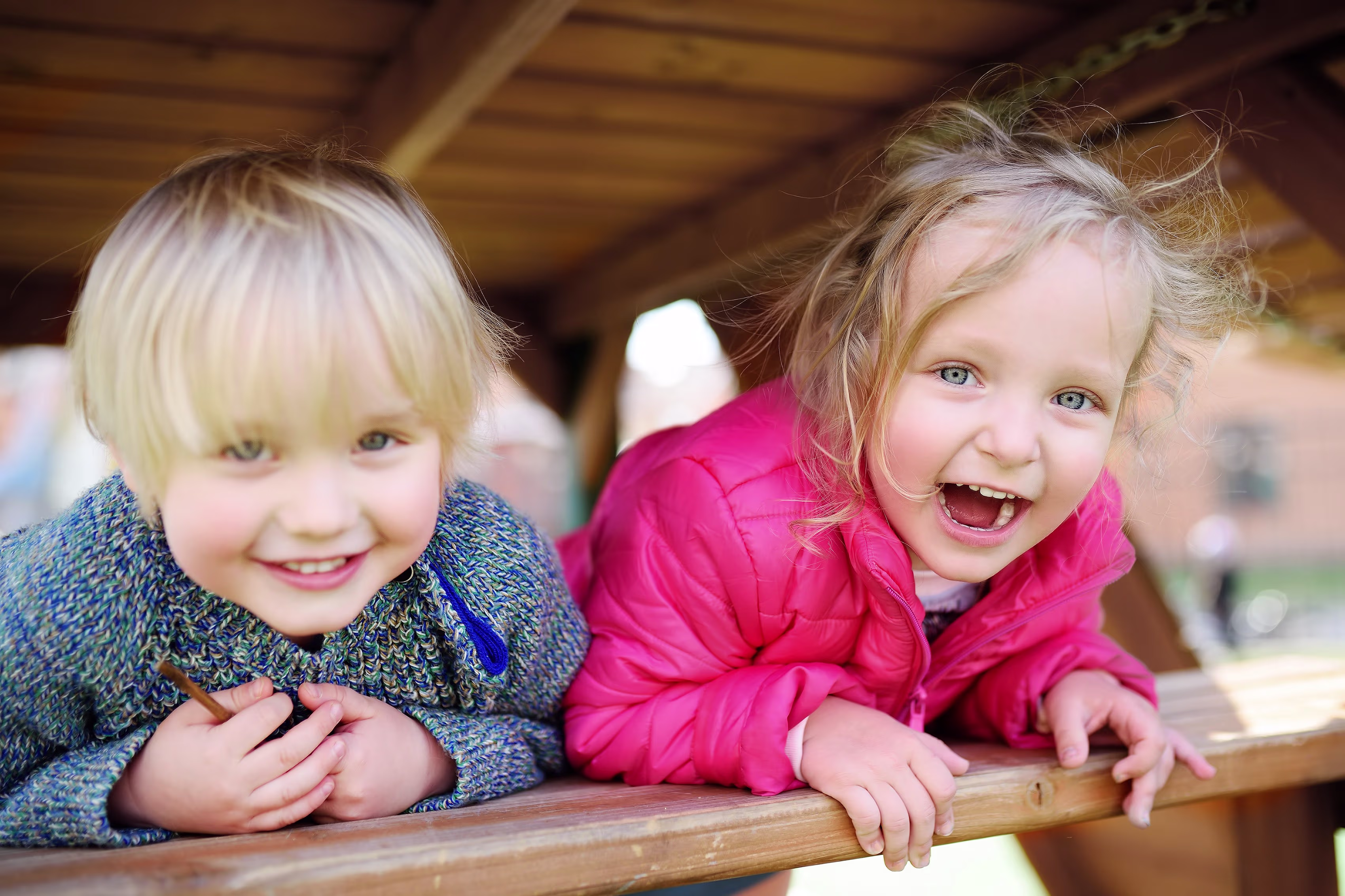 Twee blonde kinderen lachen vrolijk terwijl ze over een houten balustrade leunen in een speelomgeving.