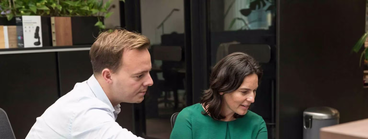 Twee collega's in gesprek achter een bureau in een modern kantoor met planten en glazen wanden op de achtergrond.