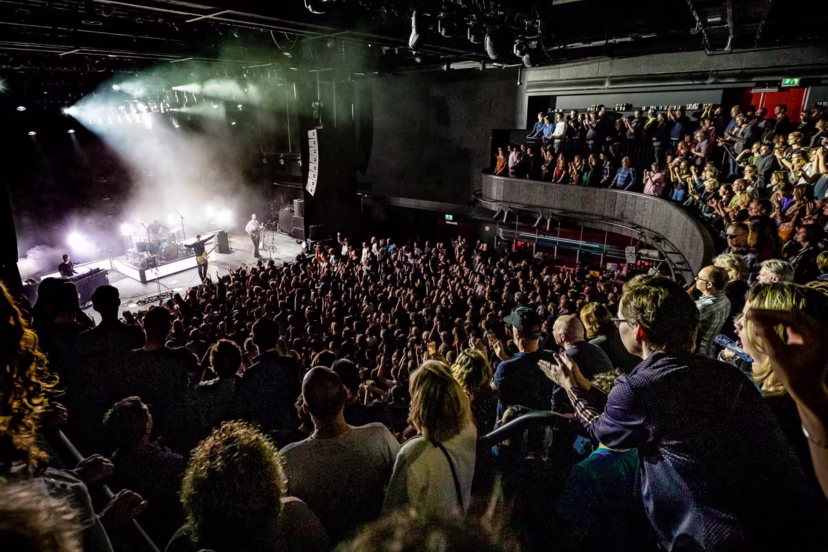 Een vol concert met publiek in een ronde concertzaal, artiest op podium met dramatische belichting en rookeffecten.
