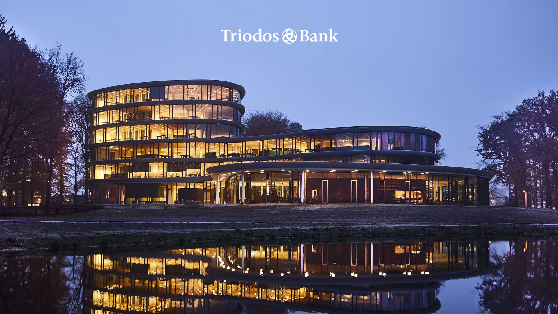 Het moderne, verlichte Triodos Bank hoofdkantoor met ronde vormen weerspiegelt in het water tijdens de schemering.