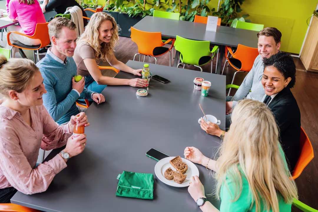 Groep collega's zit gezellig samen aan tafel in moderne kantine tijdens pauze met drankjes en snacks.