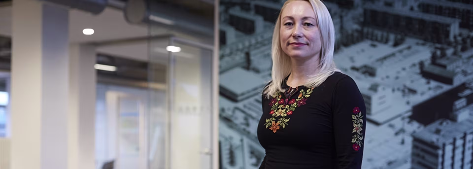 Blonde vrouw in zwarte gebloemde trui staat glimlachend in moderne kantooromgeving met glazen wanden.