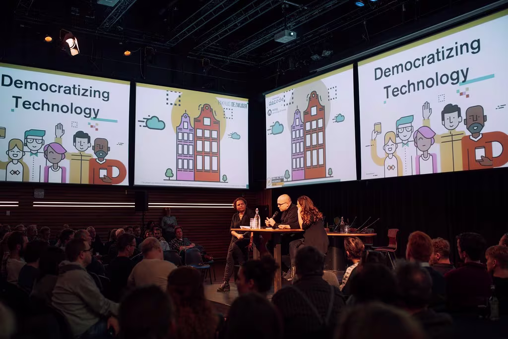 Panel discussie over "Democratizing Technology" met sprekers op podium en publiek in auditorium met grote schermen.
