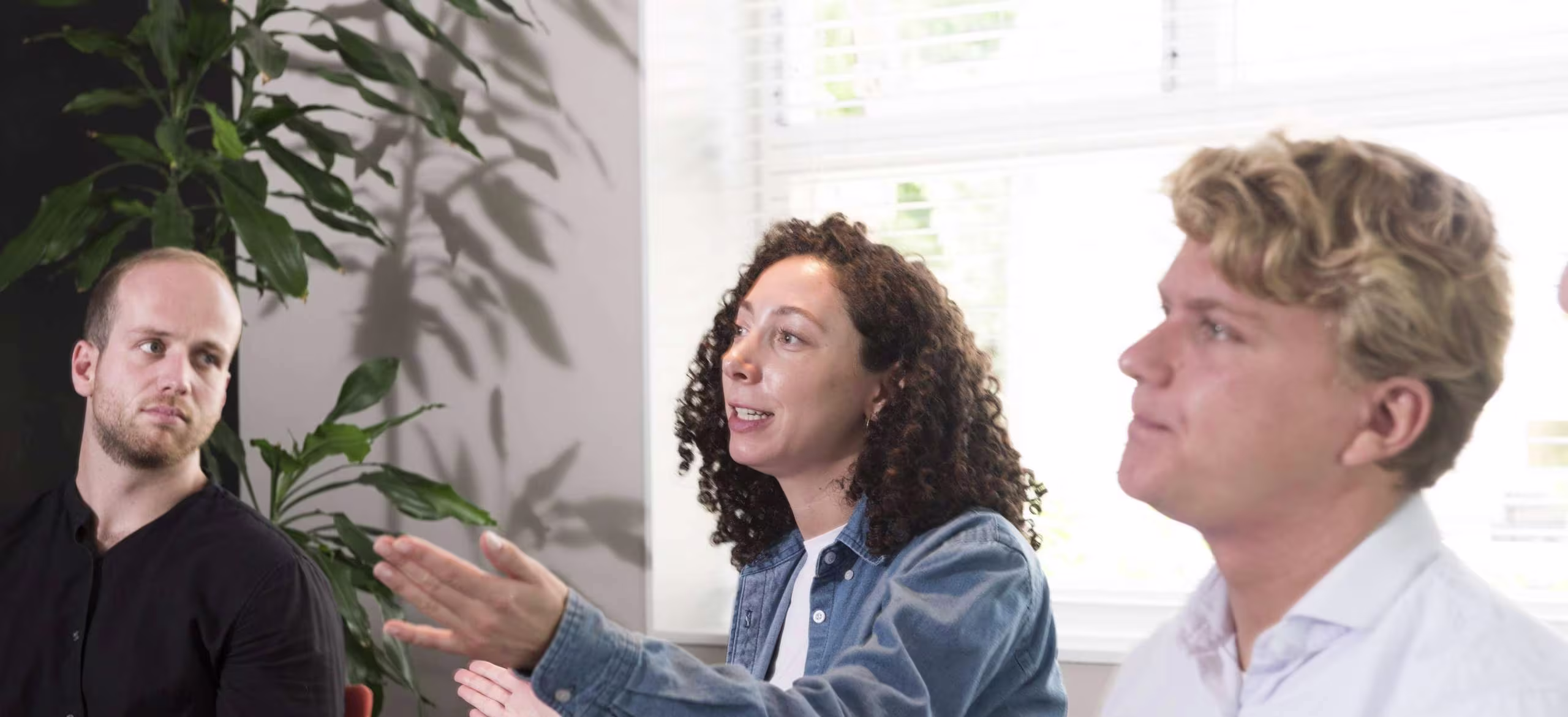 Drie collega's in gesprek op kantoor, vrouw in denim overhemd presenteert aan twee mannen bij raam met planten.