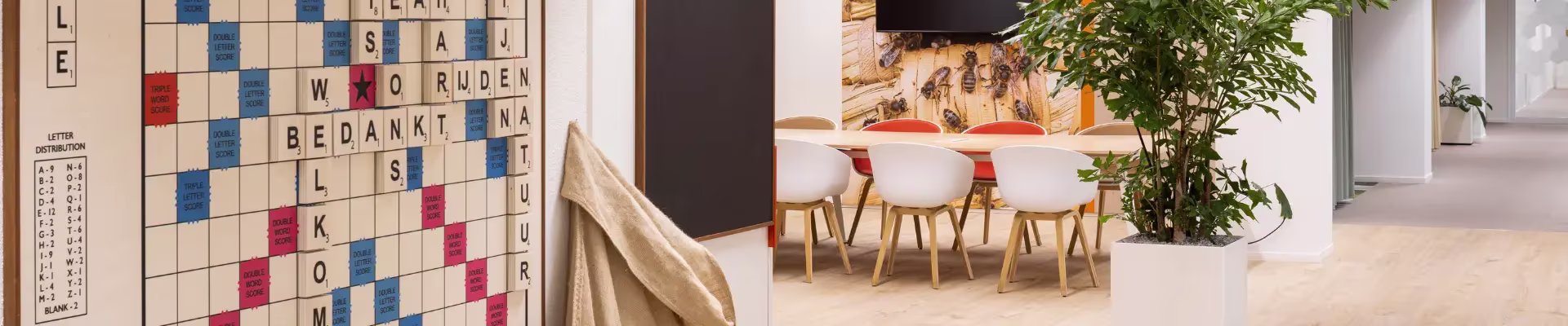 Een grote Scrabble-puzzel aan de muur naast een moderne werkruimte met witte stoelen, houten tafels en groene planten.