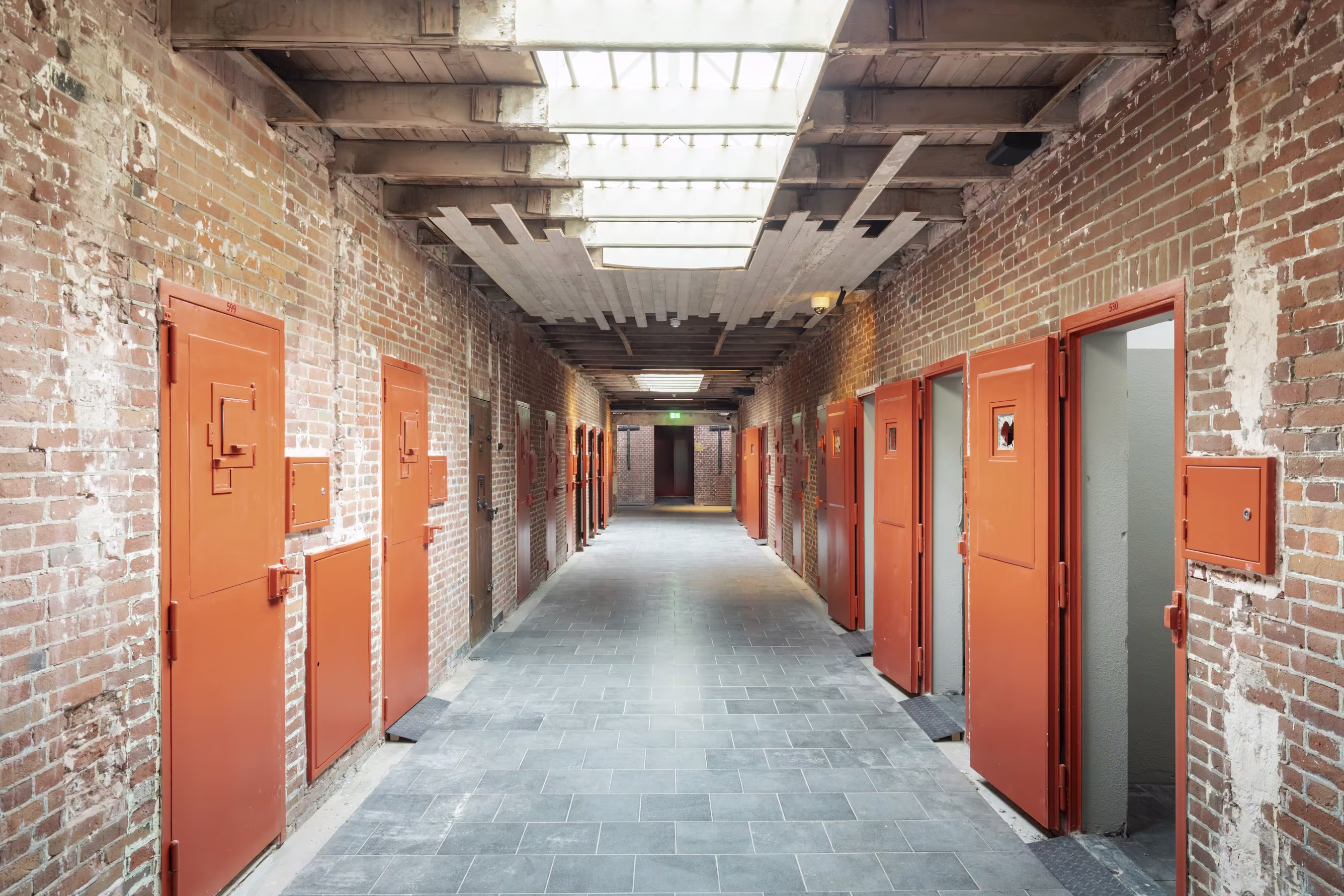 Gang met oranje deuren in historisch bakstenen gebouw met zichtbare balken en natuurlijk licht door glazen plafond.