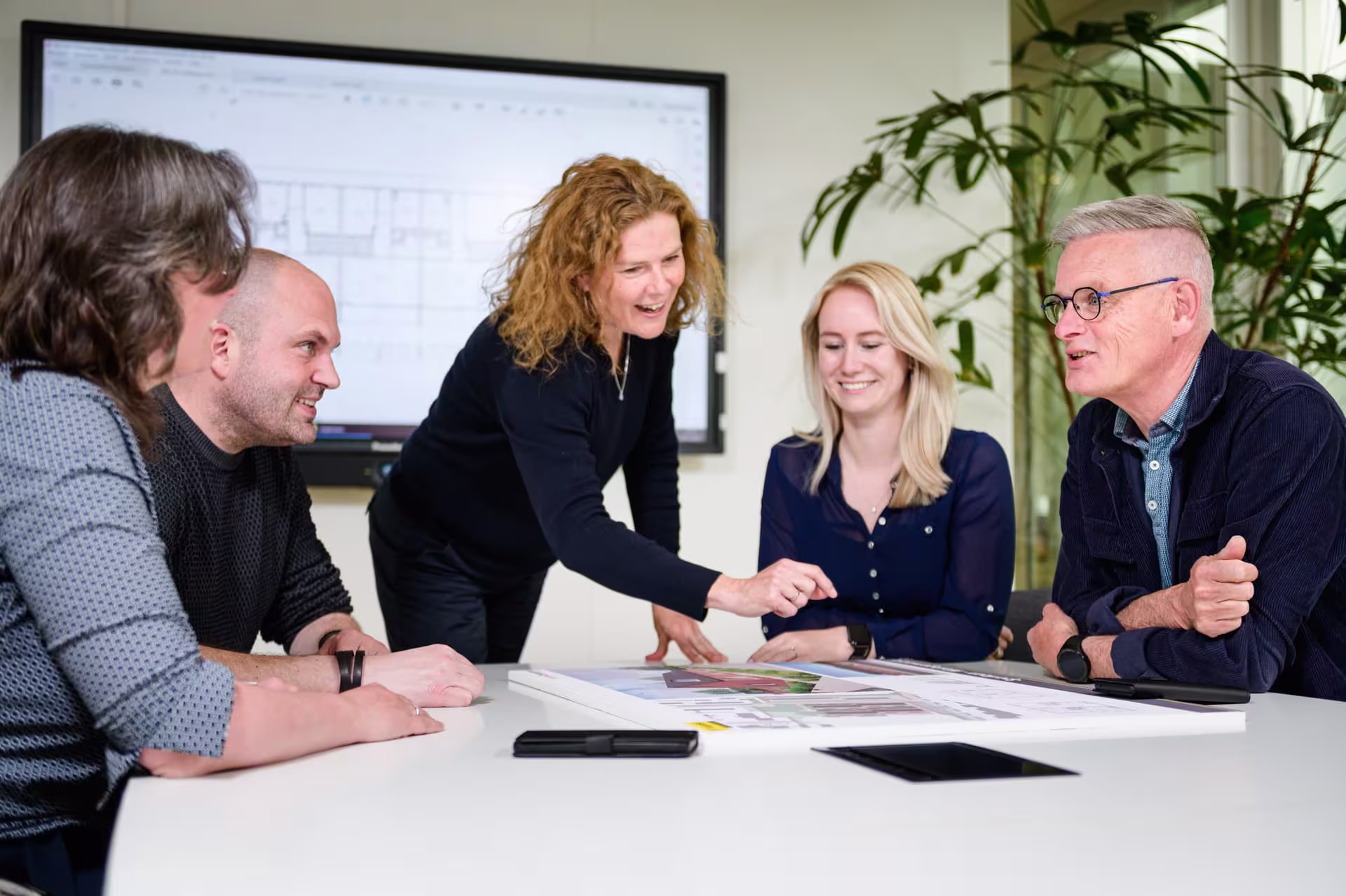 Vijf collega's bespreken samen documenten aan een vergadertafel in een moderne kantooromgeving met planten op de achtergrond.