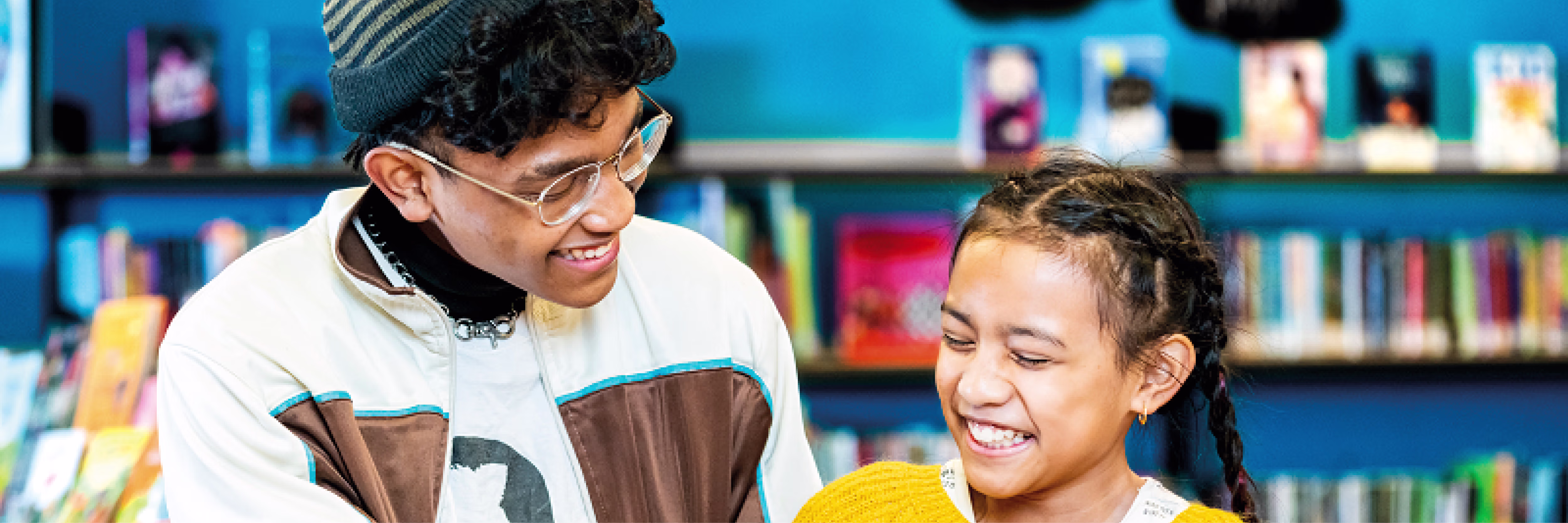Twee jongeren lachen samen terwijl ze een boek bekijken in een bibliotheek met boekenkast op de achtergrond.