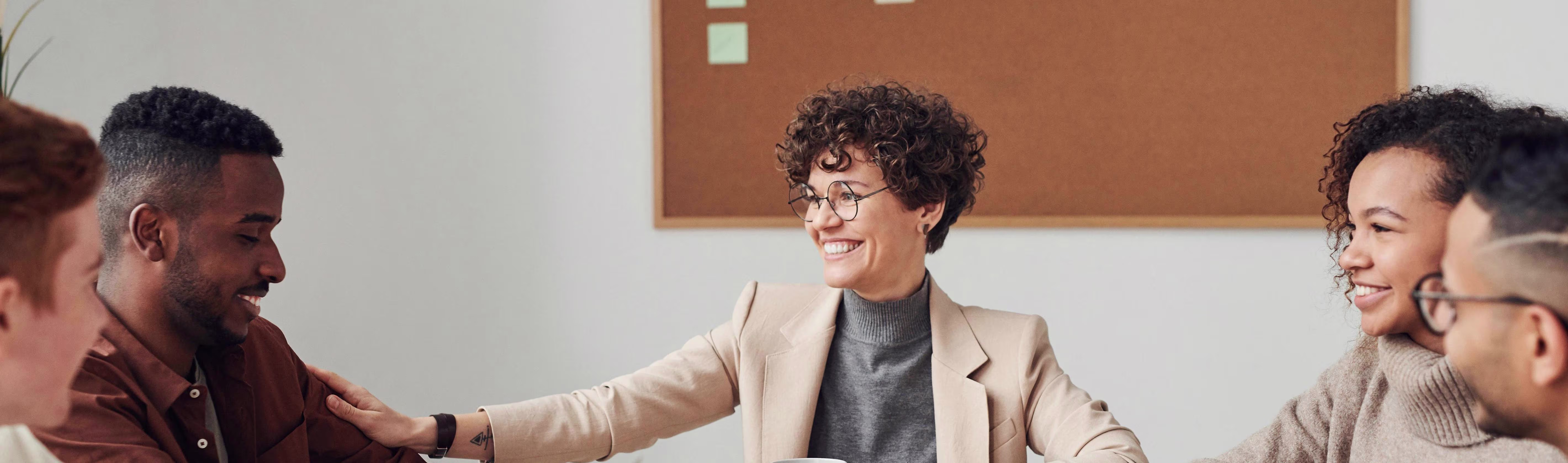 Een vrouw in beige blazer schudt handen met een man, omringd door lachende collega's in een kantooromgeving.
