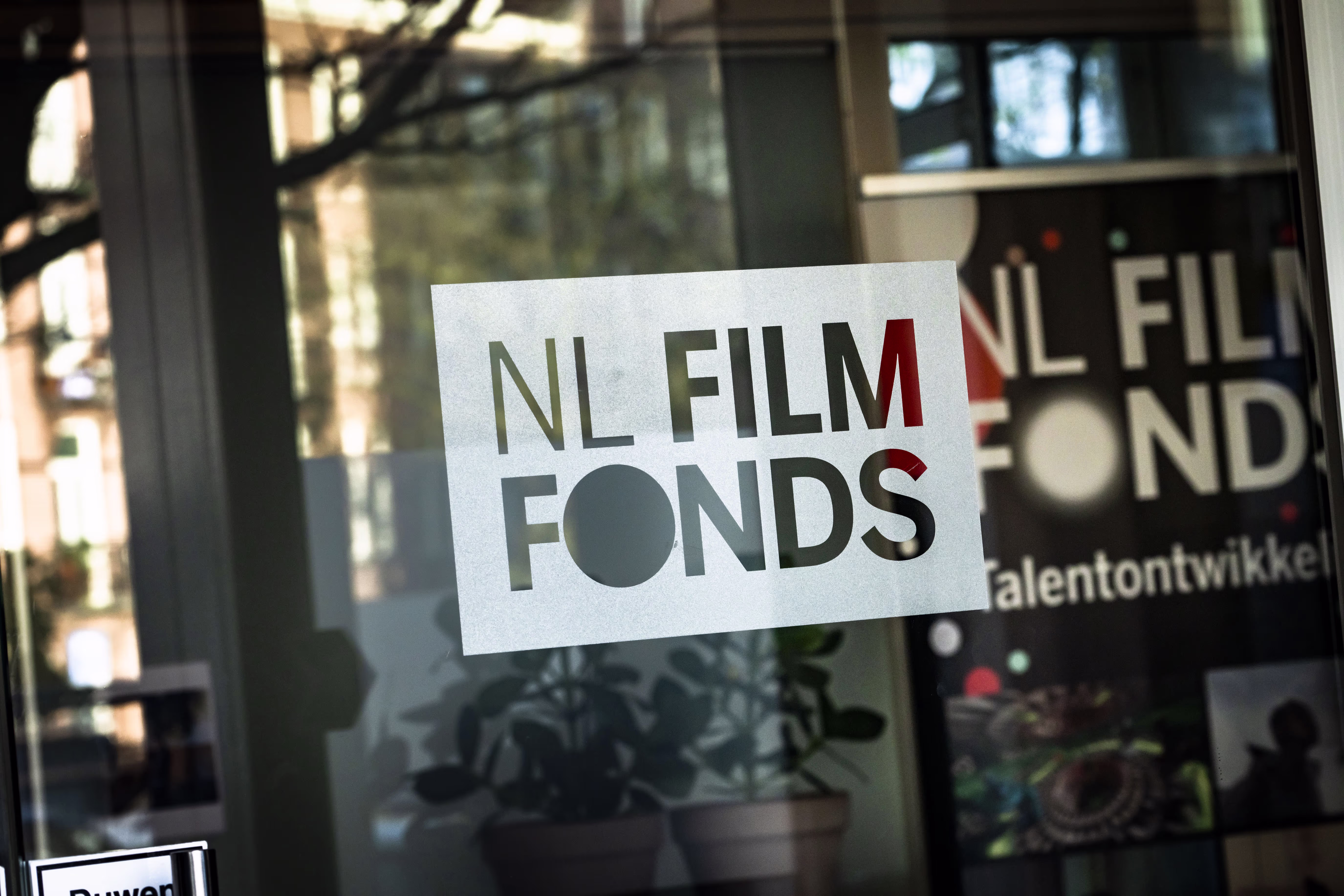 Een raam met stickers van NL Film Fonds en talentontwikkeling, met reflecties van de straat en planten zichtbaar.