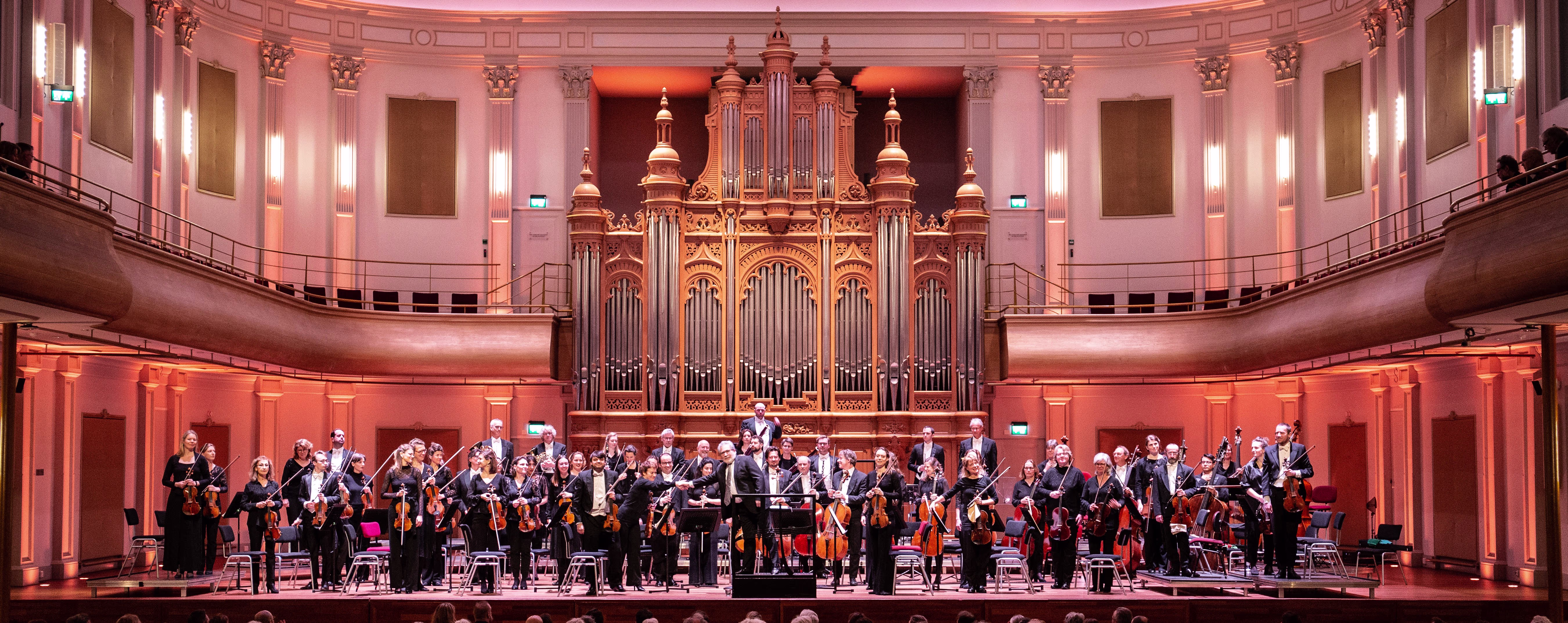 Een orkest speelt in een mooie concerthal met een groot orgel op de achtergrond en warme verlichting.