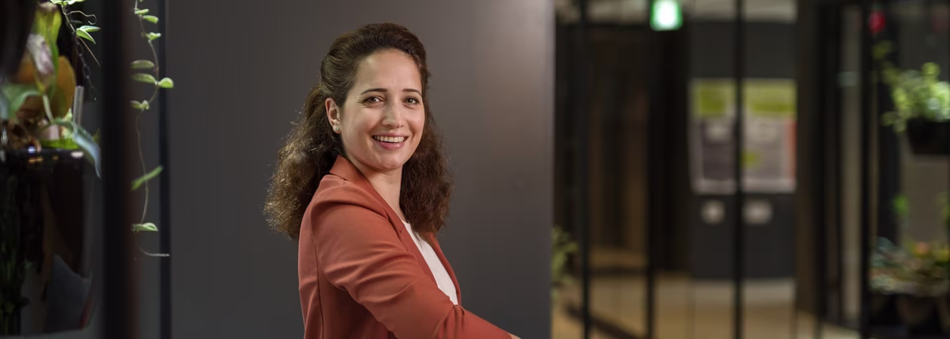 Een glimlachende vrouw in een oranje blazer draait zich om naar de camera in een modern kantoor met planten en glazen wanden.