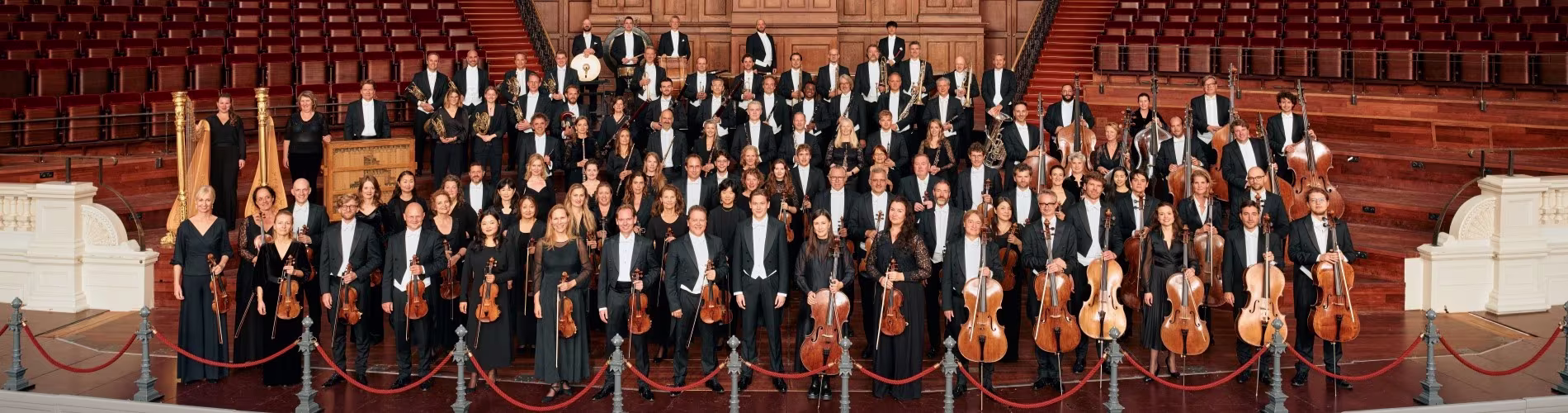 Het Koninklijk Concertgebouworkest poseert in concertzaal met strijkers vooraan en koor achteraan in formele kleding.