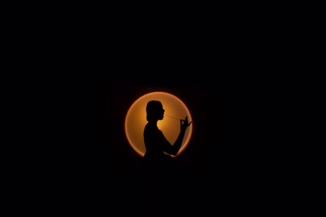 Silhouet van persoon tegen oranje/gele cirkel op zwarte achtergrond, sfeervolle artistieke compositie.