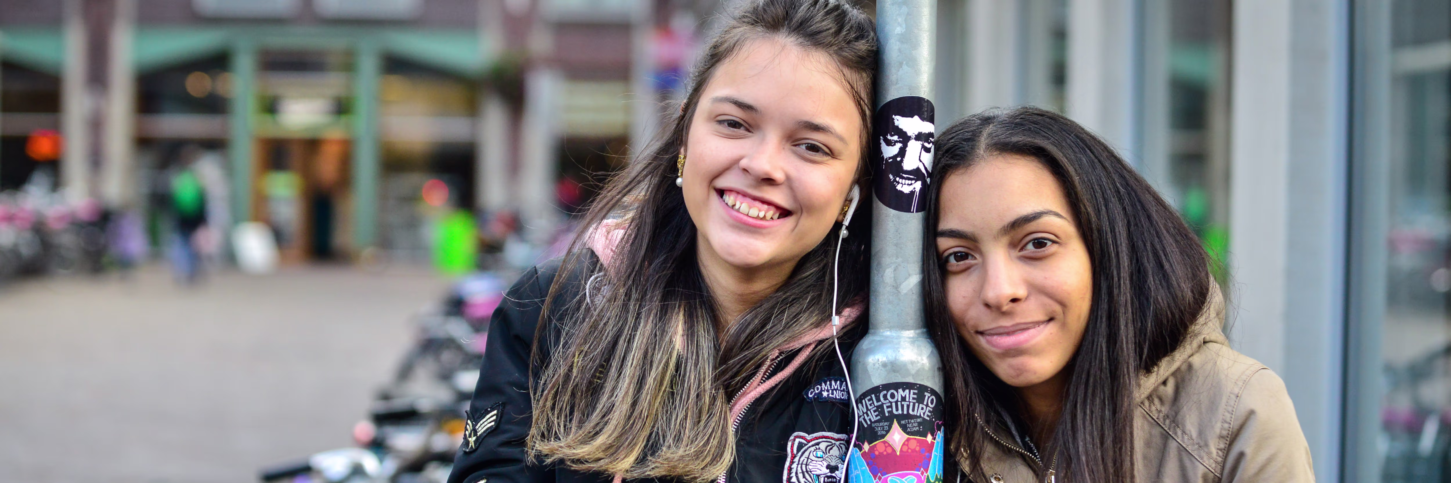 Twee lachende jonge vrouwen poseren samen met een fles op een stadsplein met winkels op de achtergrond.