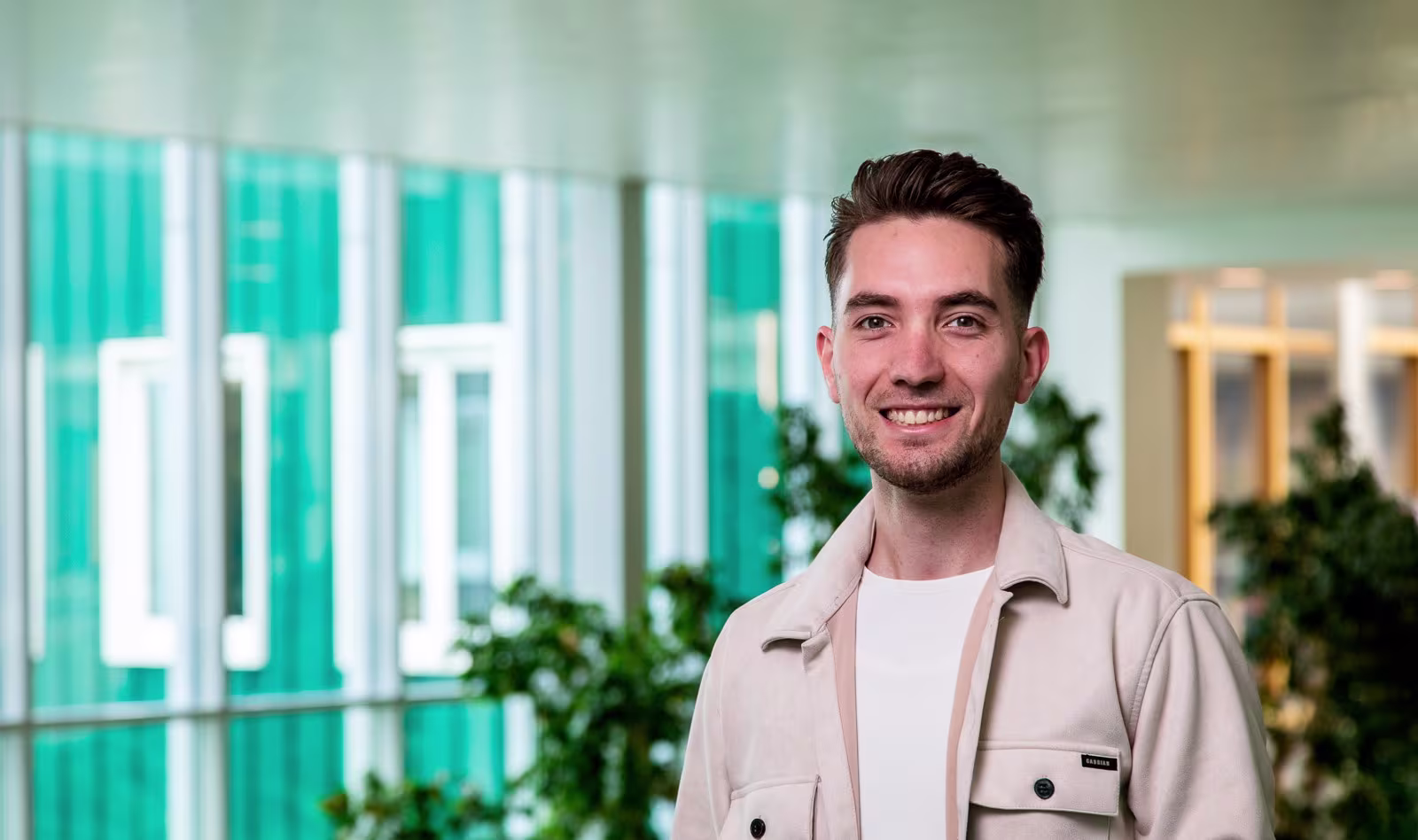 Jonge man in beige overhemd glimlacht in modern kantoorgebouw met groene planten en glazen wanden op de achtergrond.