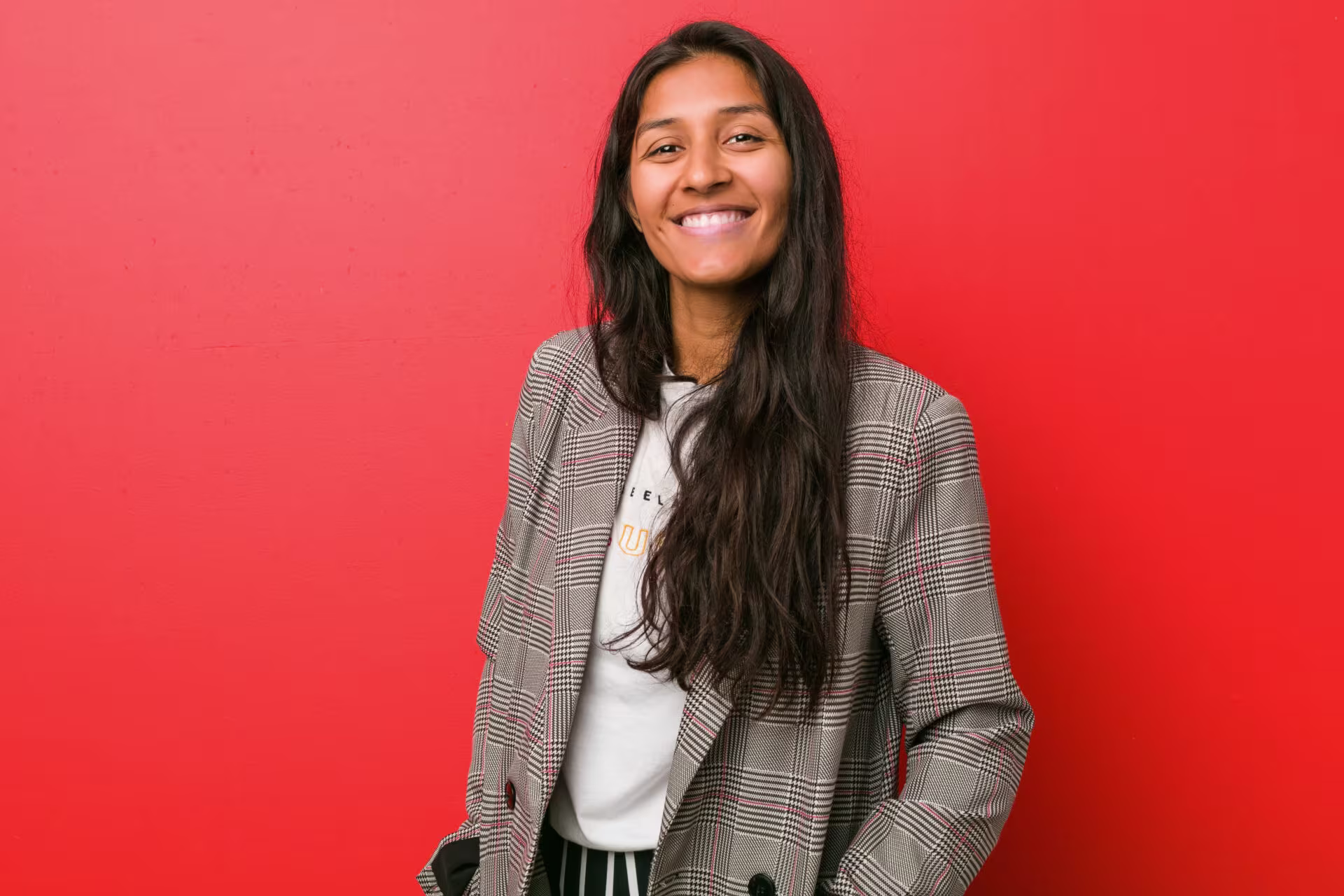 Jonge vrouw met lang donker haar lacht vriendelijk in grijze geruite blazer tegen felrode achtergrond.