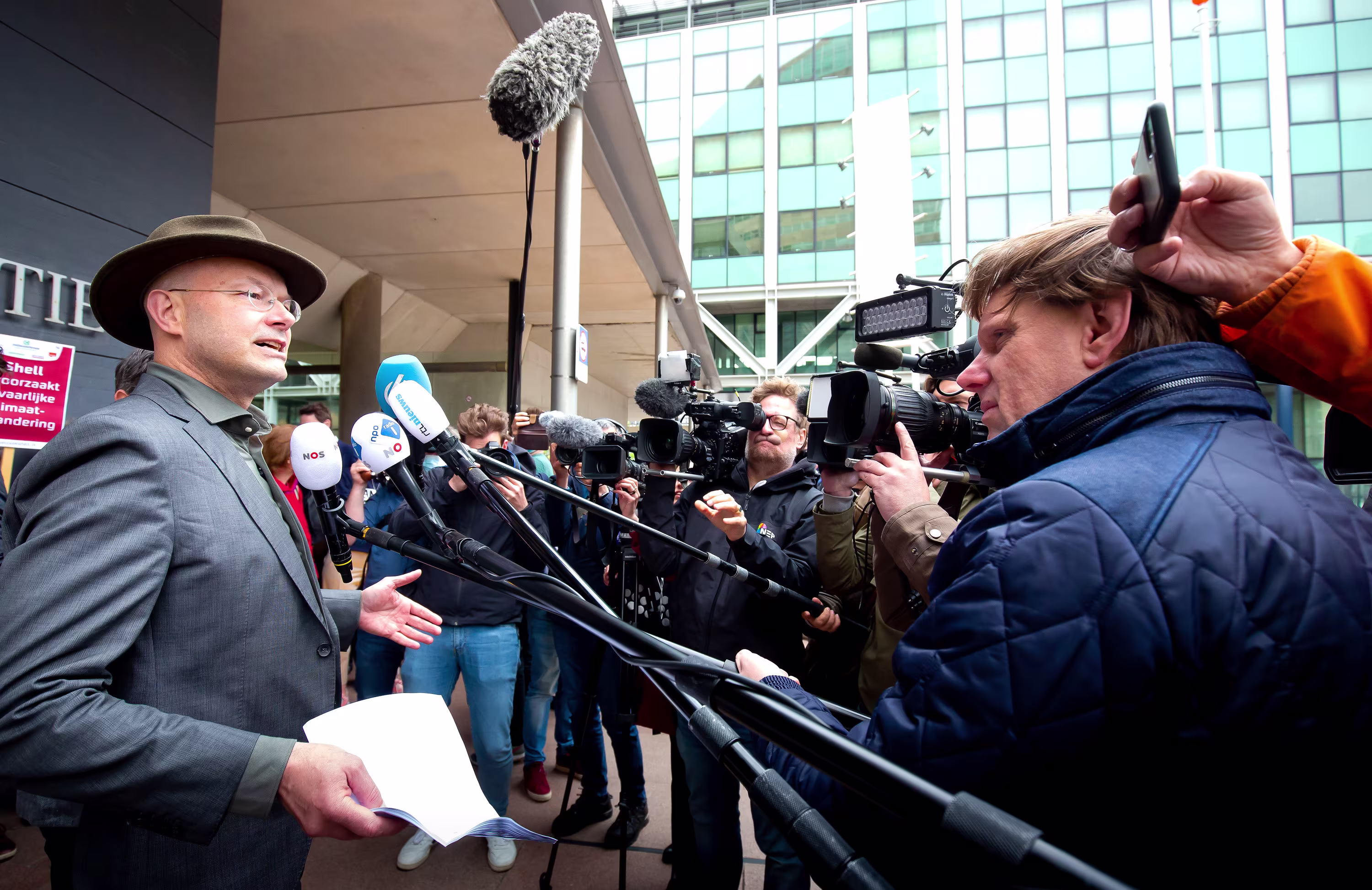 Een man in pak en hoed geeft een persconferentie met veel microfoons en journalisten met camera's om hem heen.