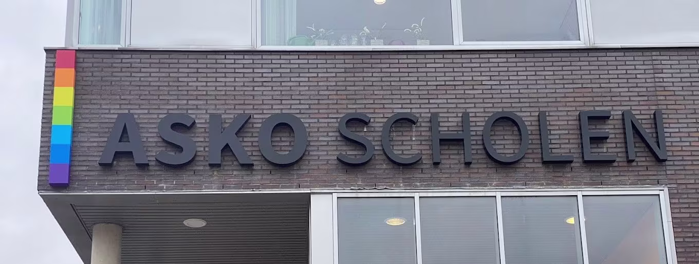 Gevel van ASKO SCHOLEN met regenboogstrepen en grote letters op bakstenen muur, glazen ramen zichtbaar.