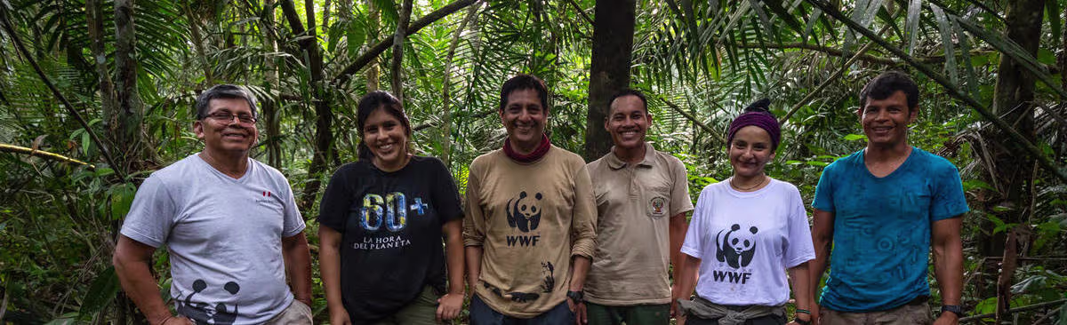 Zes medewerkers van WWF staan lachend in een tropisch regenwoud, gekleed in WWF-shirts en werkkleding.