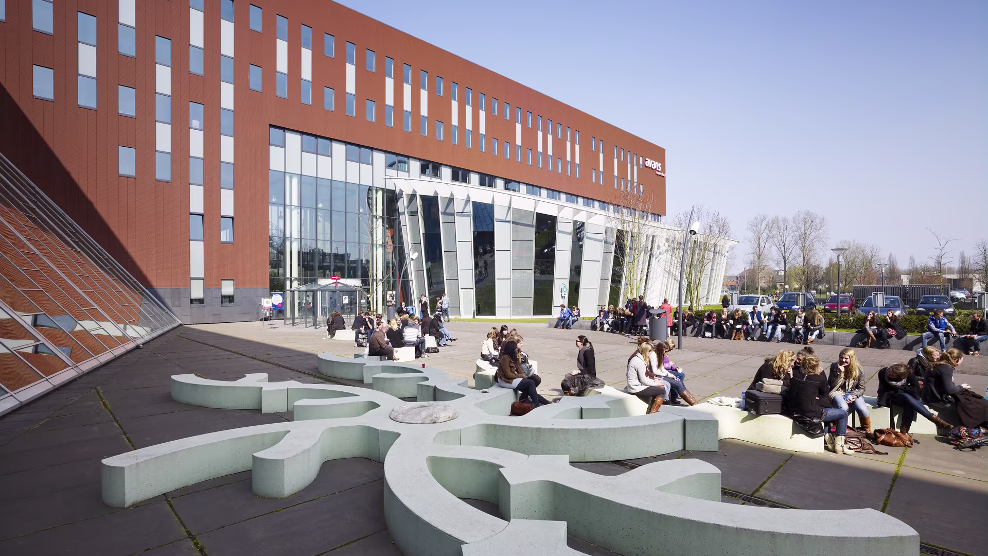Studenten zitten buiten op het plein voor het moderne Avans Hogeschool gebouw met rode gevel en grote glazen entree.