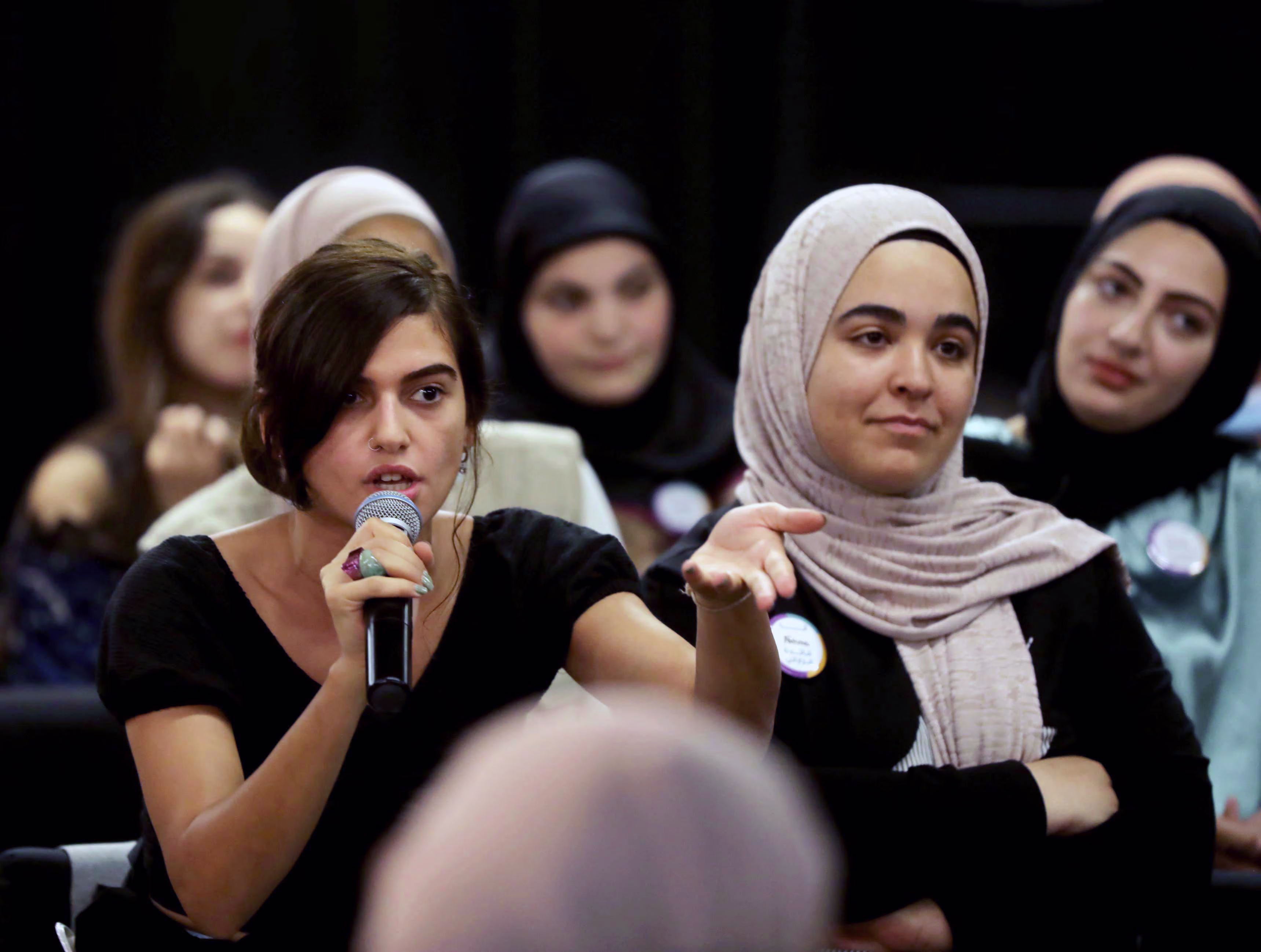Vrouw met microfoon spreekt op evenement, naast haar zit vrouw met hoofddoek, publiek op achtergrond.