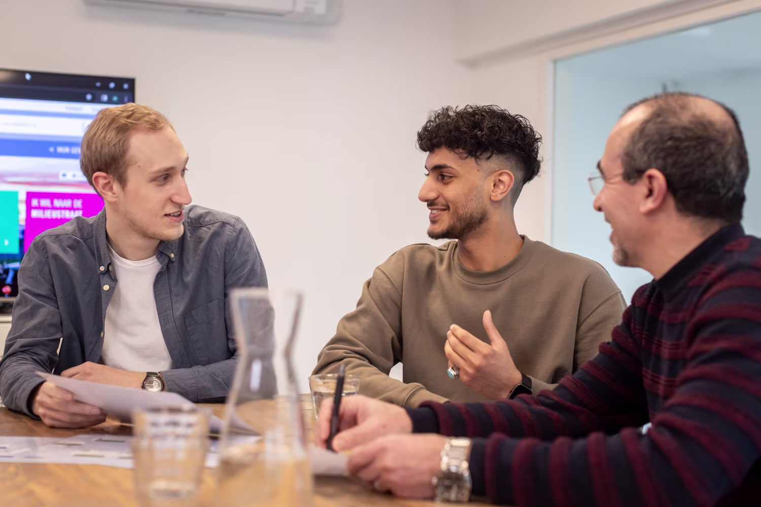 Drie mannen zitten aan een tafel in gesprek in een moderne kantooromgeving met computer op de achtergrond.