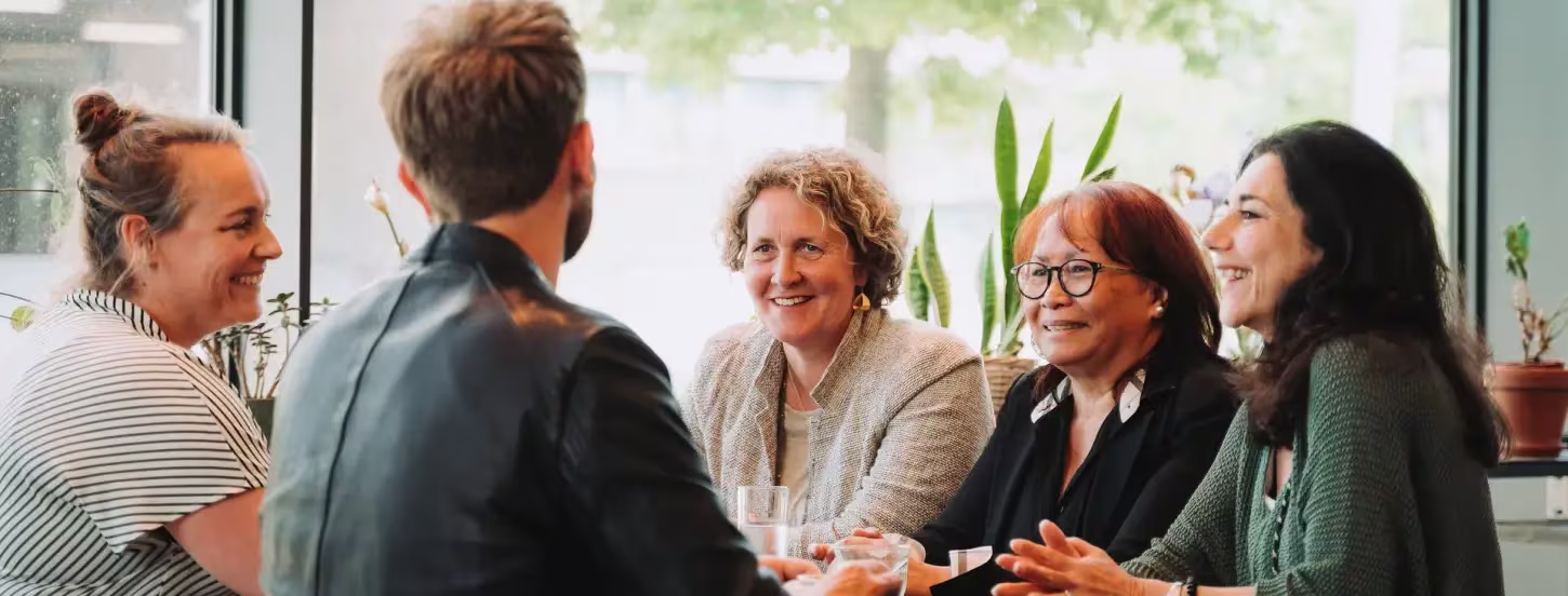 Groep collega's in gesprek rond tafel bij raam, lachend en geanimeerd discussiërend in kantooromgeving.