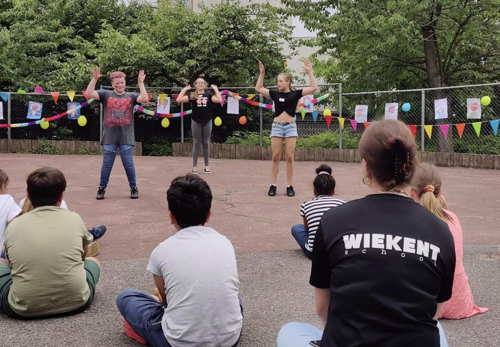 Drie vrouwen geven een buitenoptreden voor een zittend publiek, met kleurrijke slingers op de achtergrond.