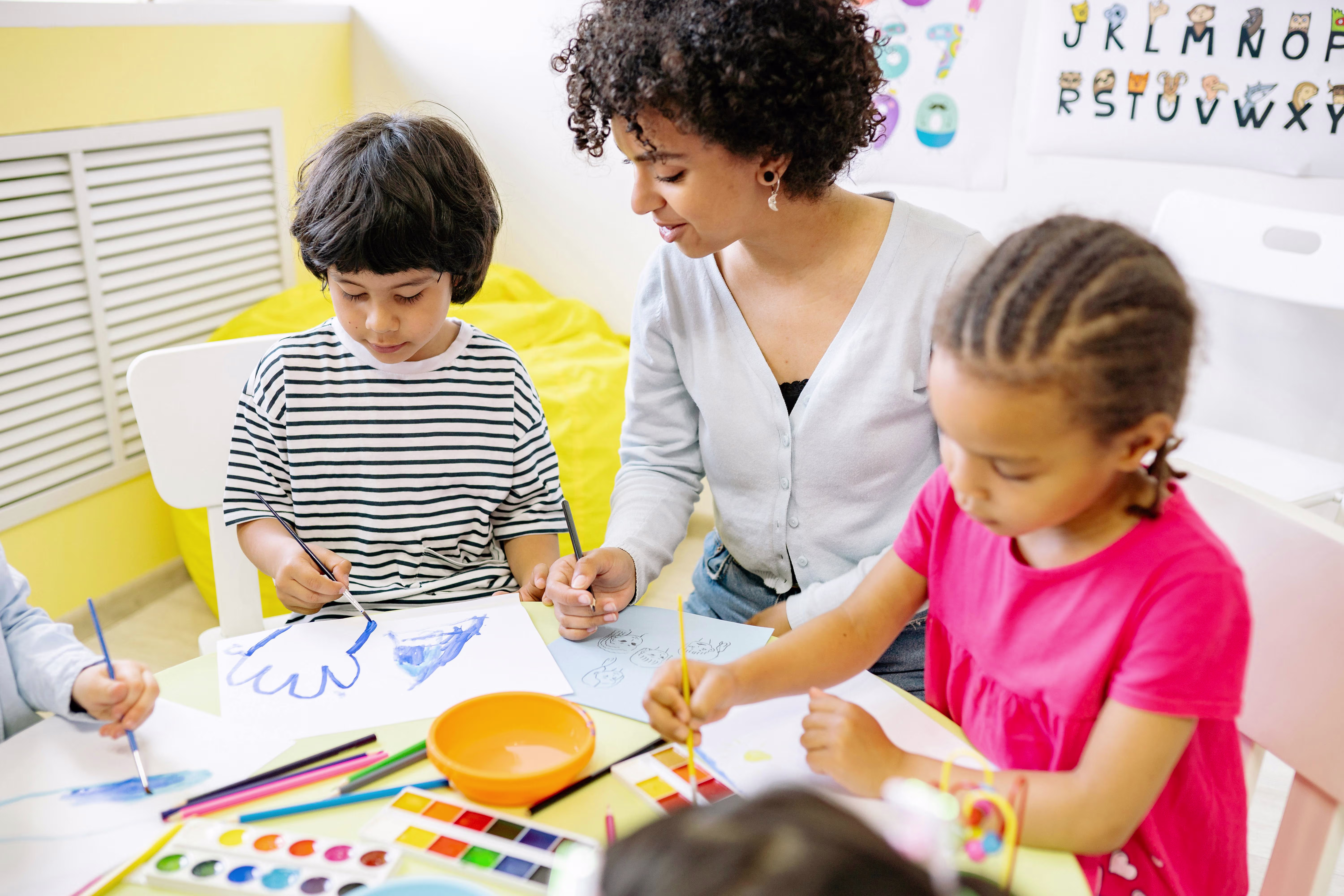 Een onderwijsmedewerker begeleidt drie kinderen tijdens een creatieve tekenactiviteit in de klas met verfpaletten en papier.