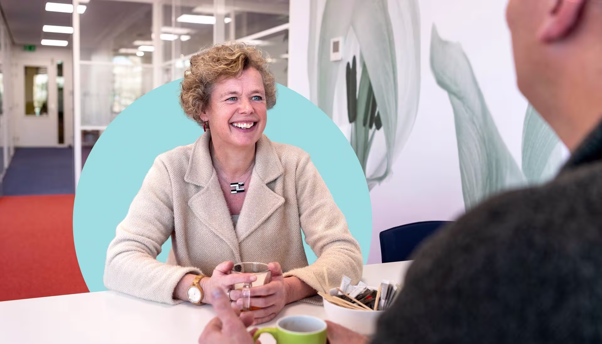 Lachende vrouw in beige blazer in gesprek aan tafel in modern kantoor van Slachtofferhulp Nederland.