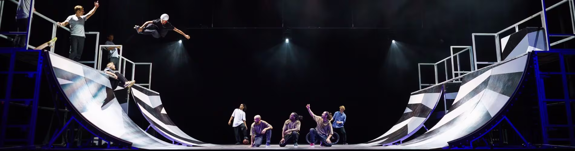 Dansers presteren op een moderne theaterset met gebogen platforms en dramatische verlichting tijdens een voorstelling.