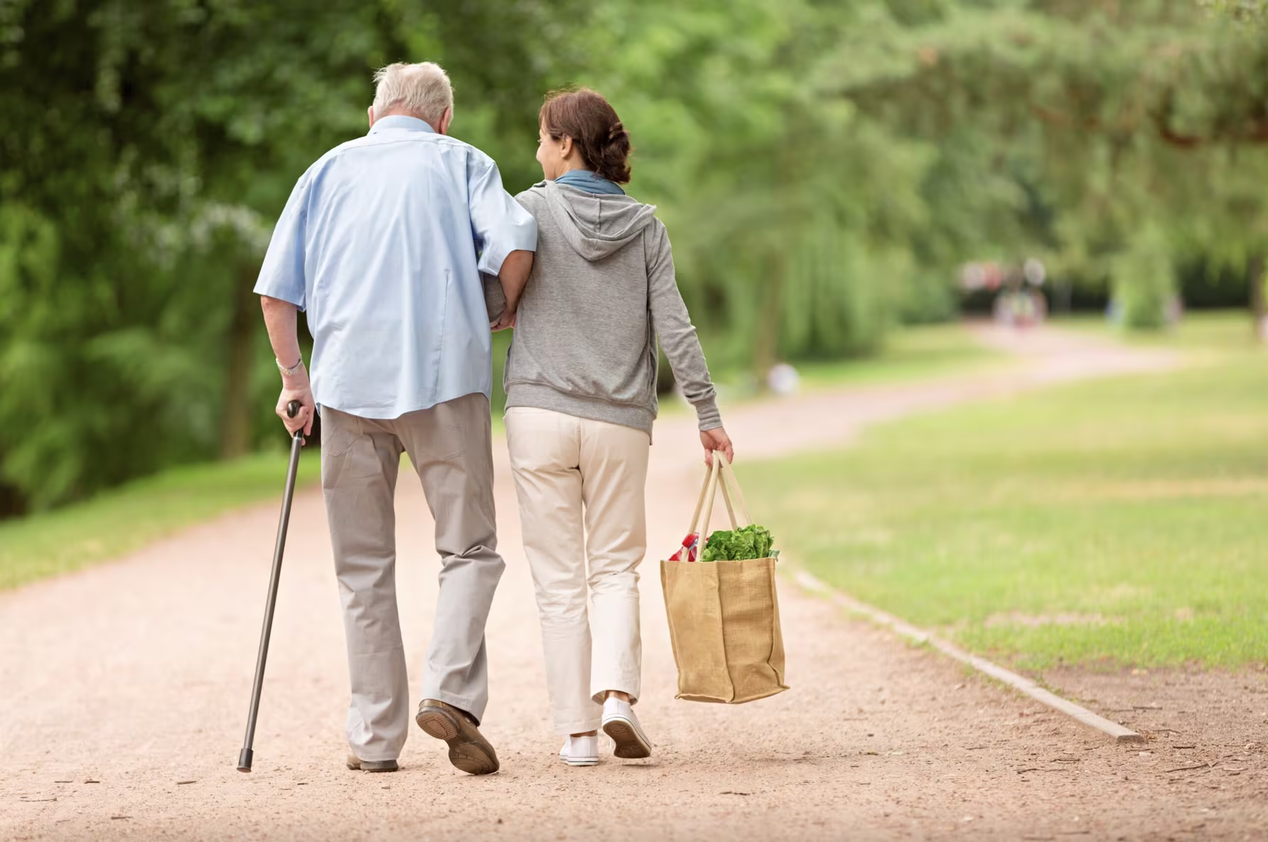 Een oudere man met wandelstok en een vrouw lopen samen over een parkpad, zij draagt een boodschappentas.