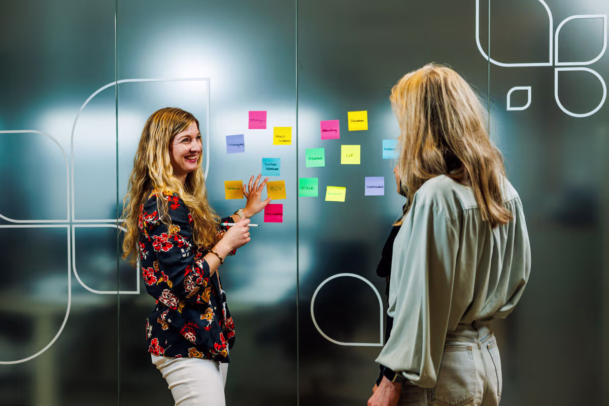 Twee vrouwen bespreken kleurrijke post-it notities op een glazen wand tijdens een brainstormsessie op kantoor.