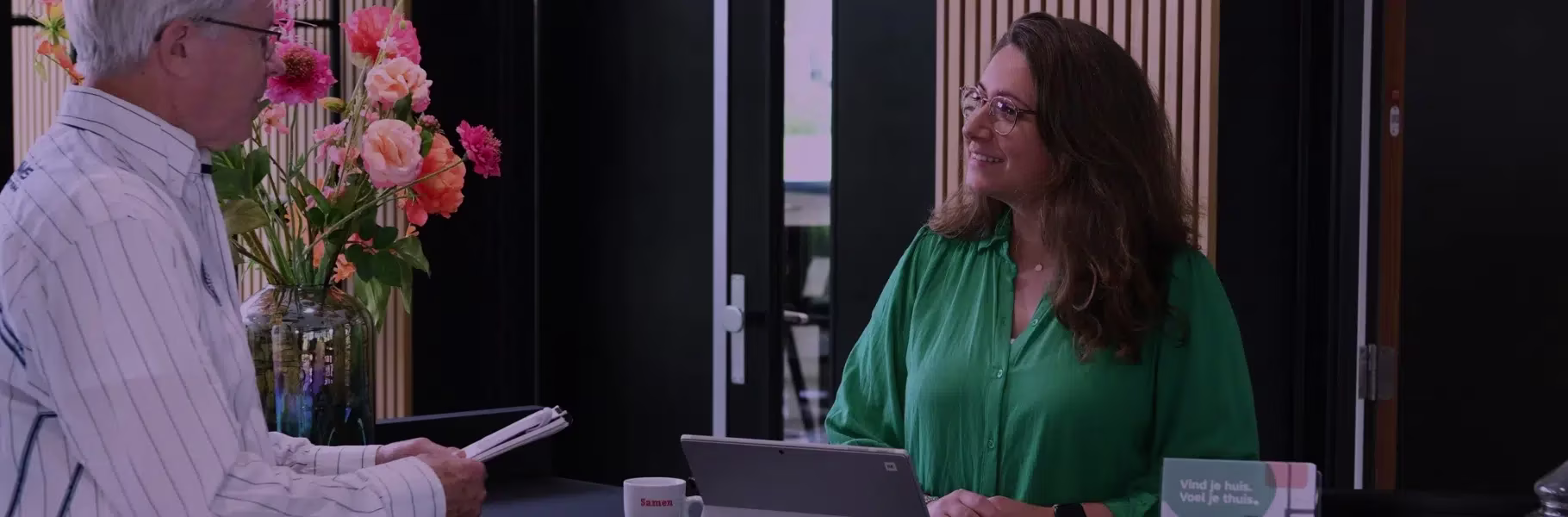 Een man en vrouw in gesprek op kantoor, zij draagt een groene blouse en zit achter een bureau met laptop en bloemen.