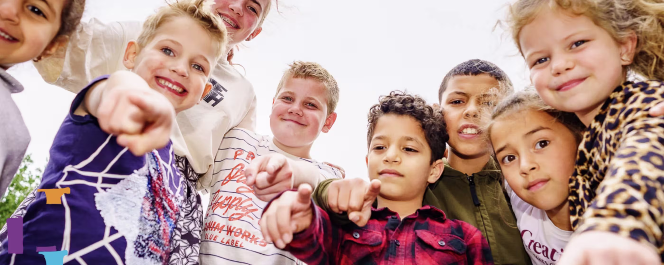 Een groep diverse kinderen steekt lachend hun handen naar de camera uit, gefotografeerd van onderaf tegen een witte hemel.