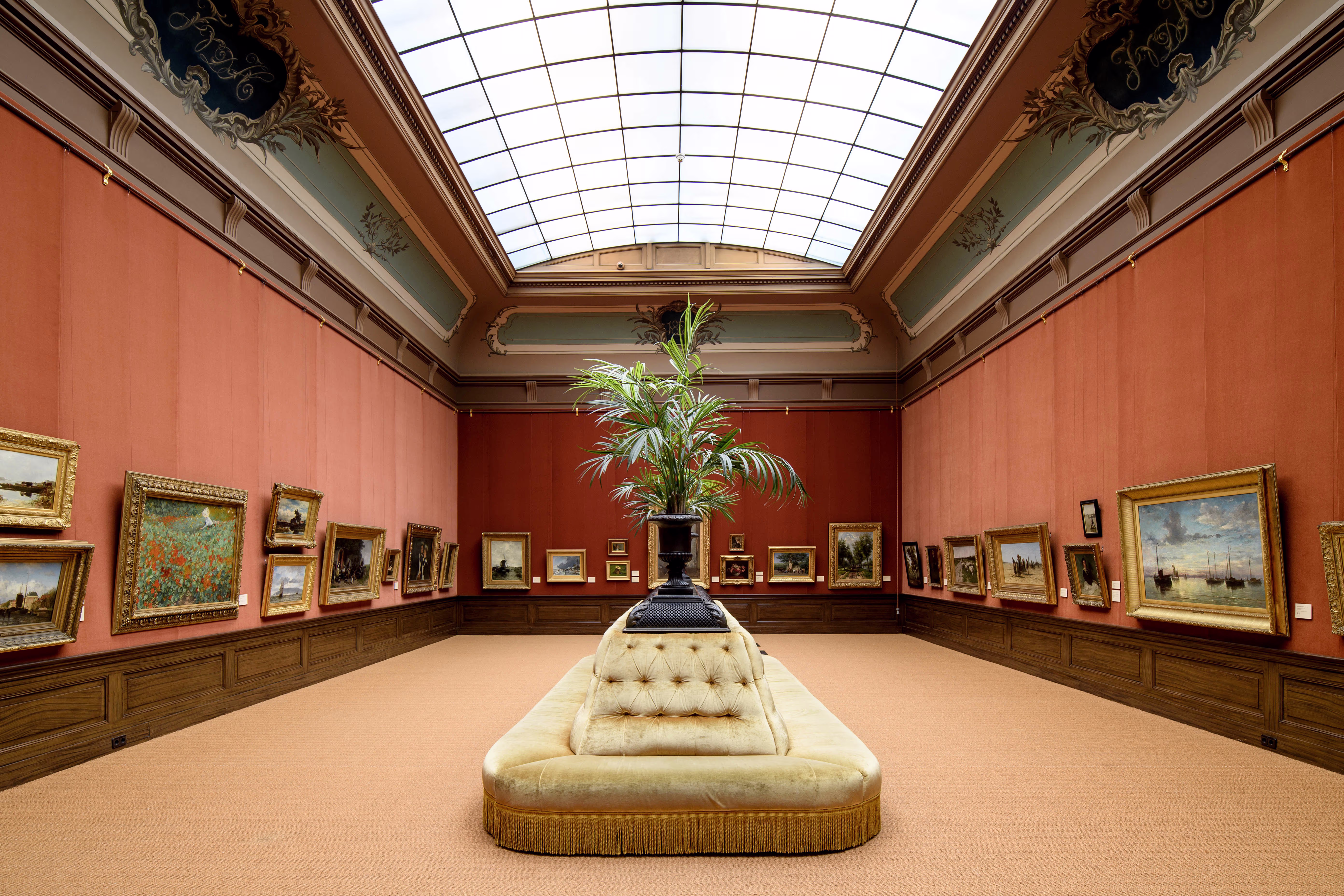 Elegante museumzaal met oranje muren, schilderijen in gouden lijsten, glazen koepeldak en een fluwelen bank in het midden.