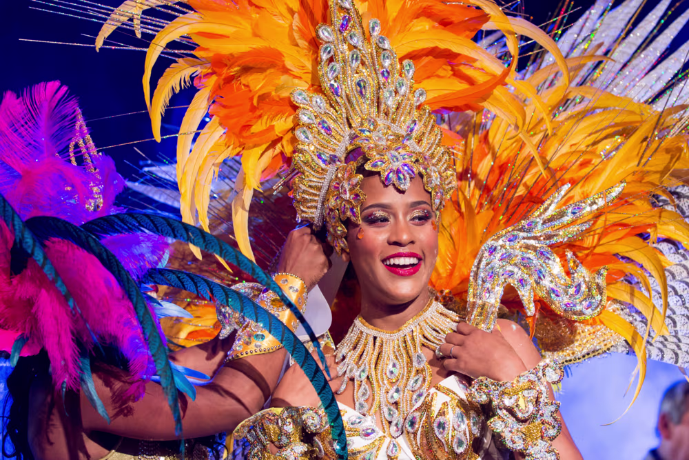 Vrouw in kleurrijk carnavalskostuum met oranje veren en gouden versieringen tijdens een feestelijk evenement.