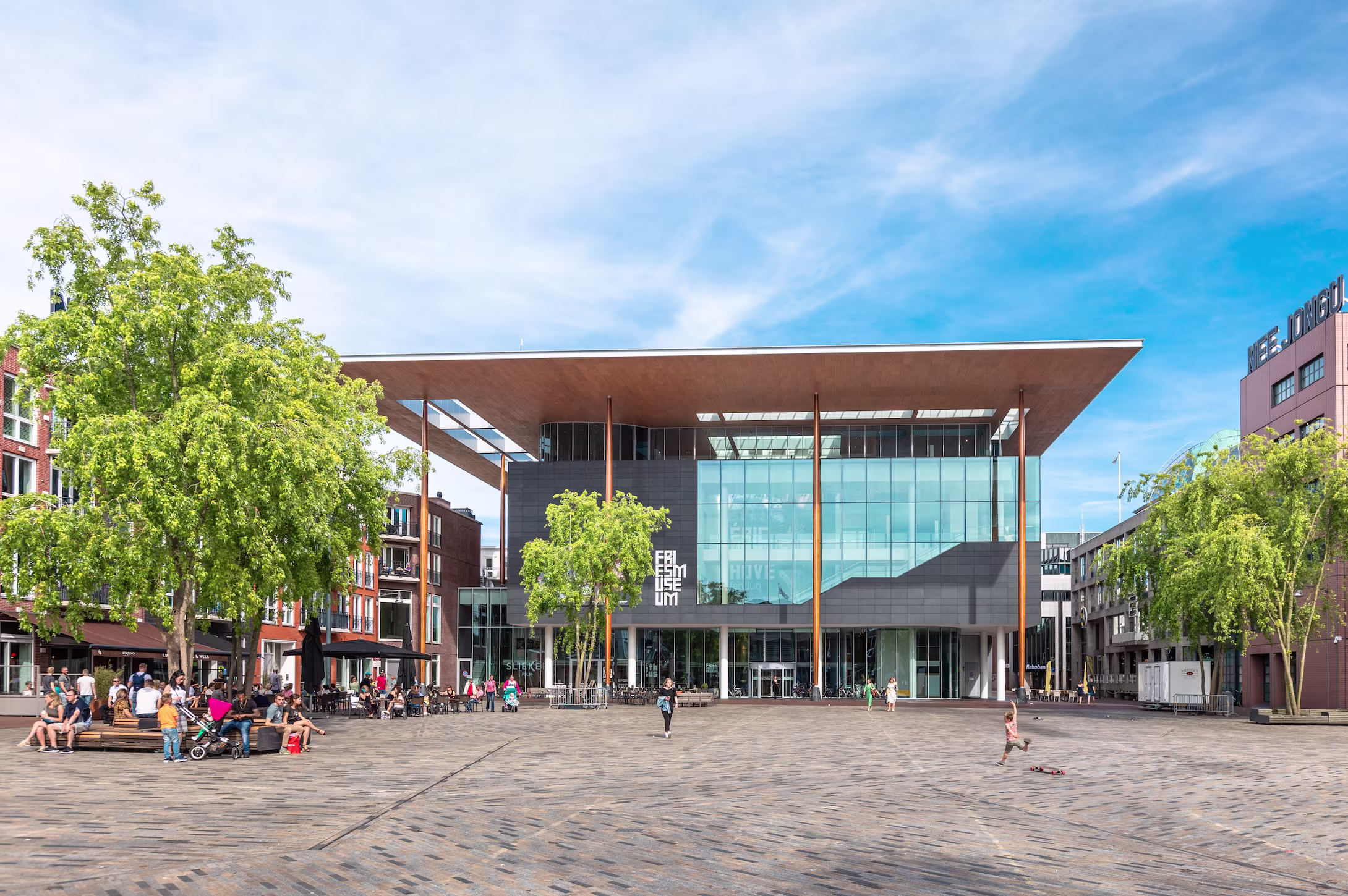 Het moderne Fries Museum gebouw met glazen gevel en groot dak op een plein met mensen en historische panden eromheen.