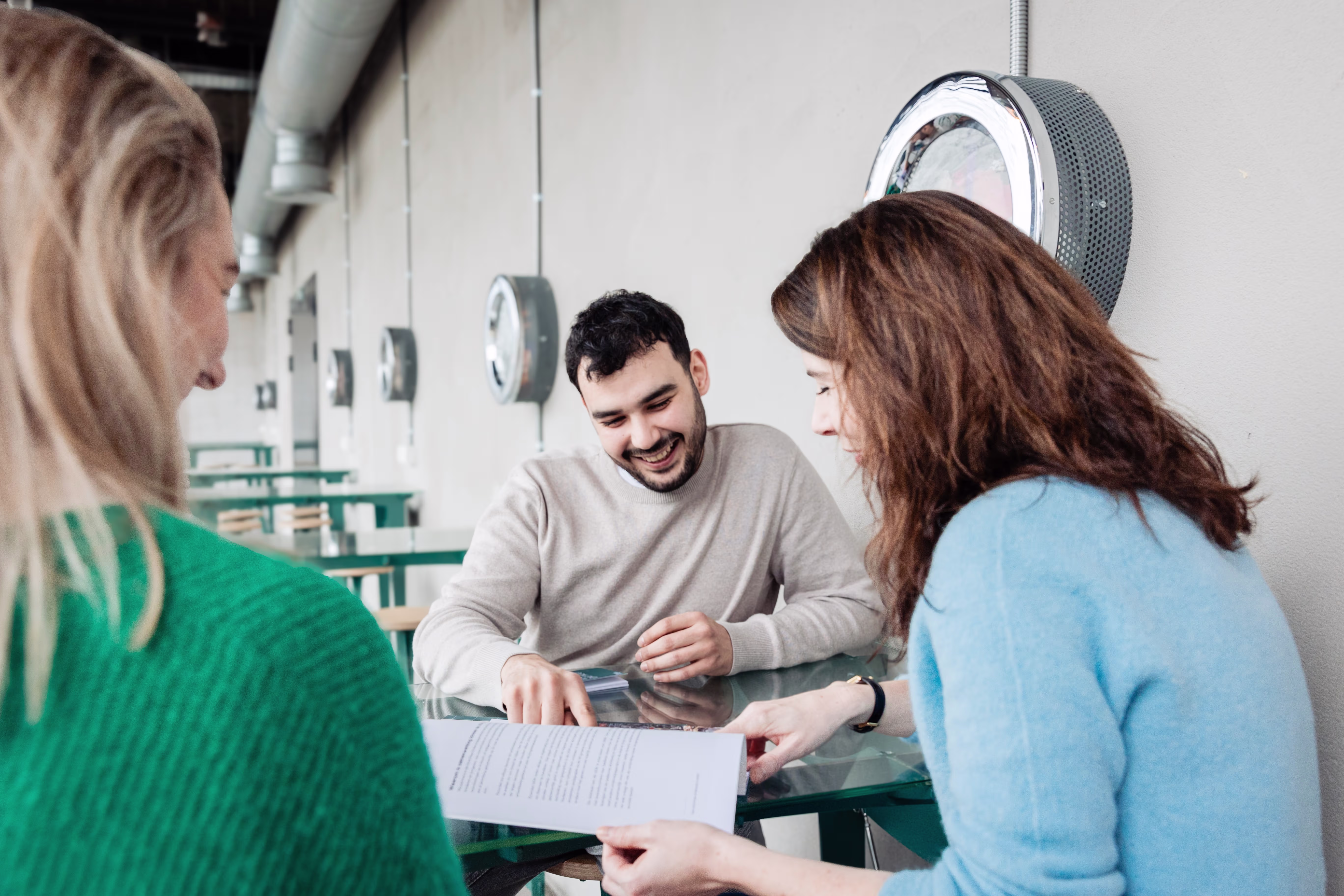Drie collega's bespreken documenten aan een tafel in een modern kantoor met ronde wandlampen op de achtergrond.