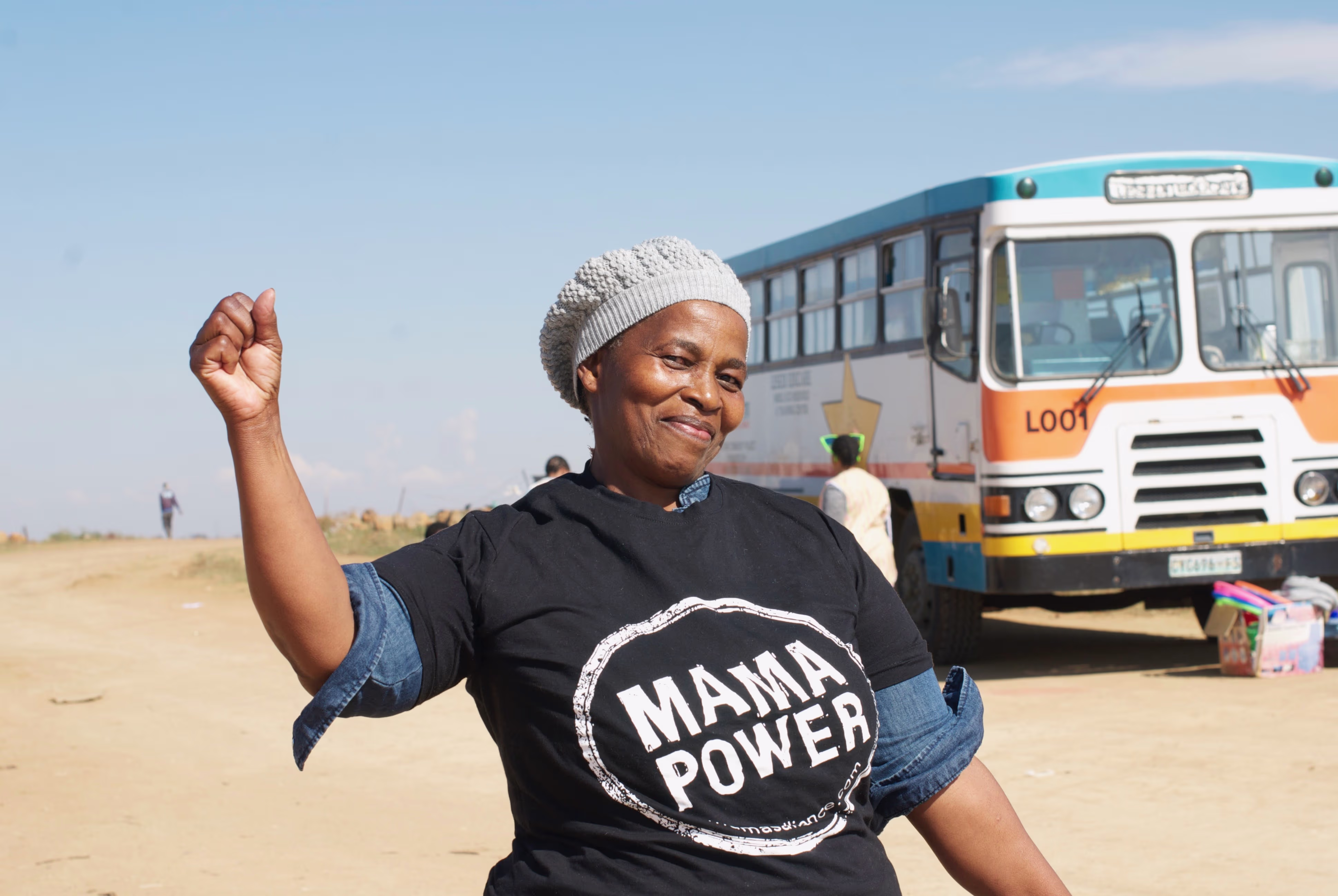 Glimlachende vrouw in zwart 'Mama Power' shirt steekt vrolijk haar hand op, met bus op achtergrond in zonnige omgeving.