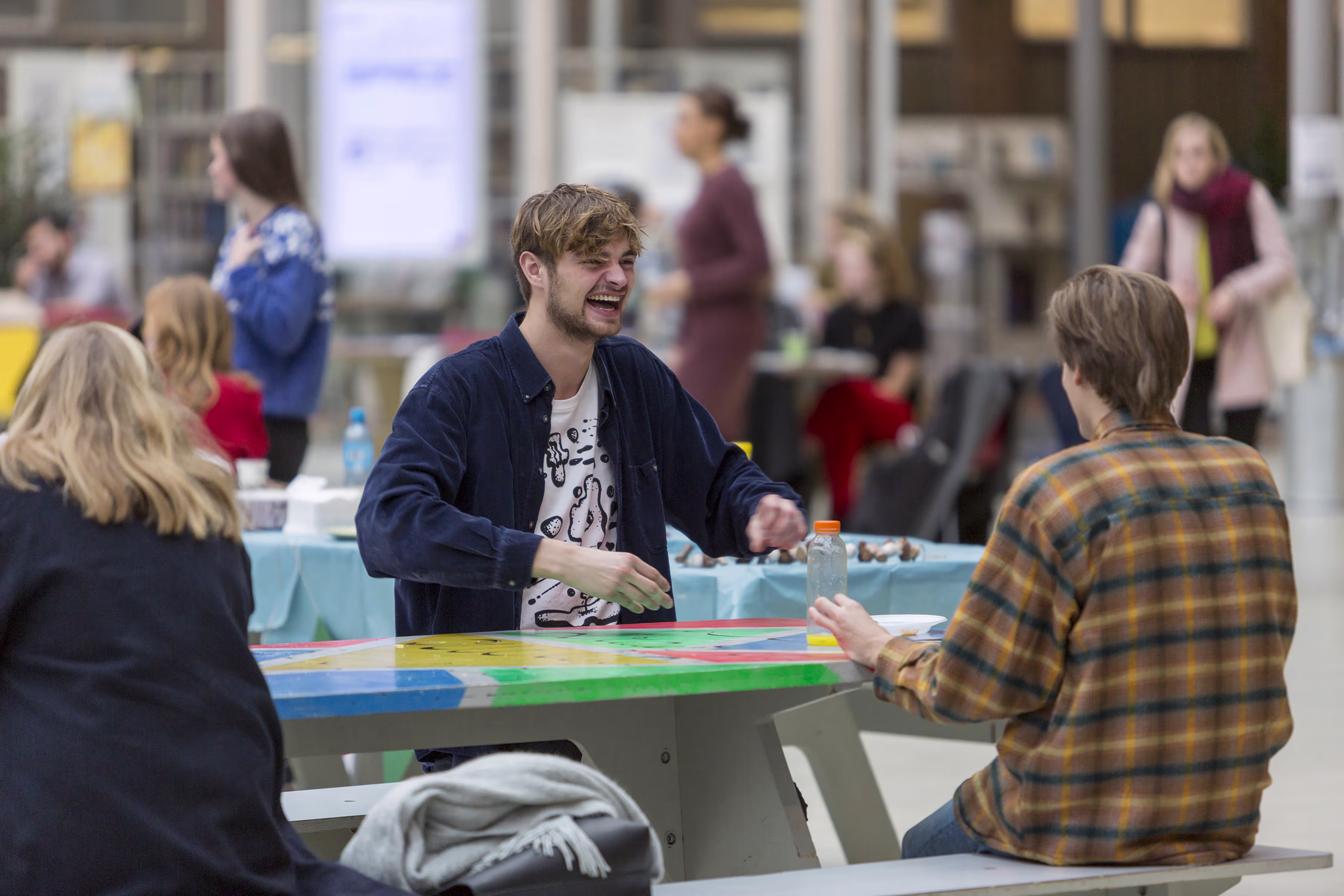 Studenten zitten gezellig samen aan een kleurrijke tafel in een moderne onderwijsruimte en praten vrolijk met elkaar.