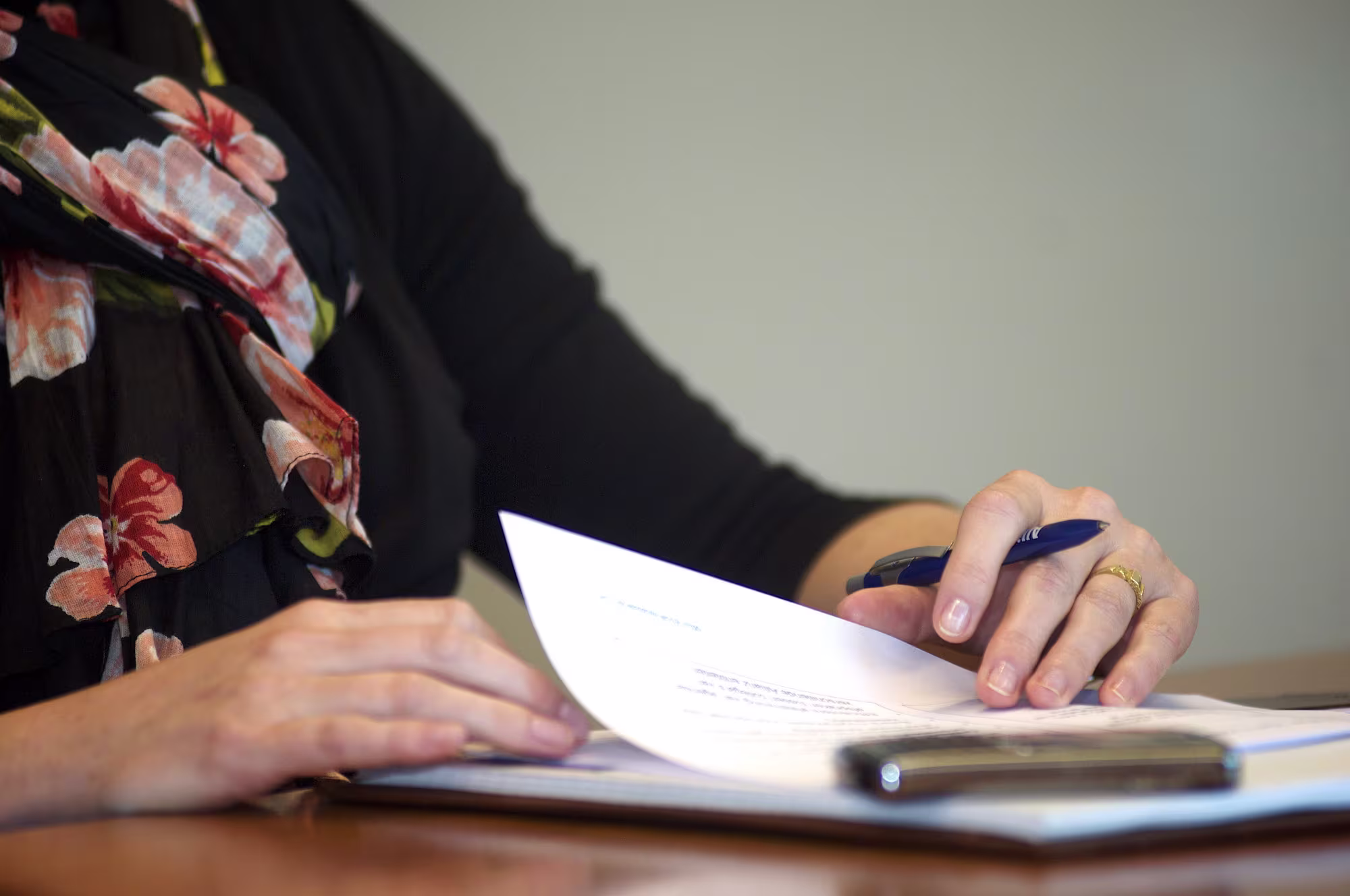 Een persoon in een zwarte blouse met bloemenprint leest documenten aan een bureau met een pen in de hand.