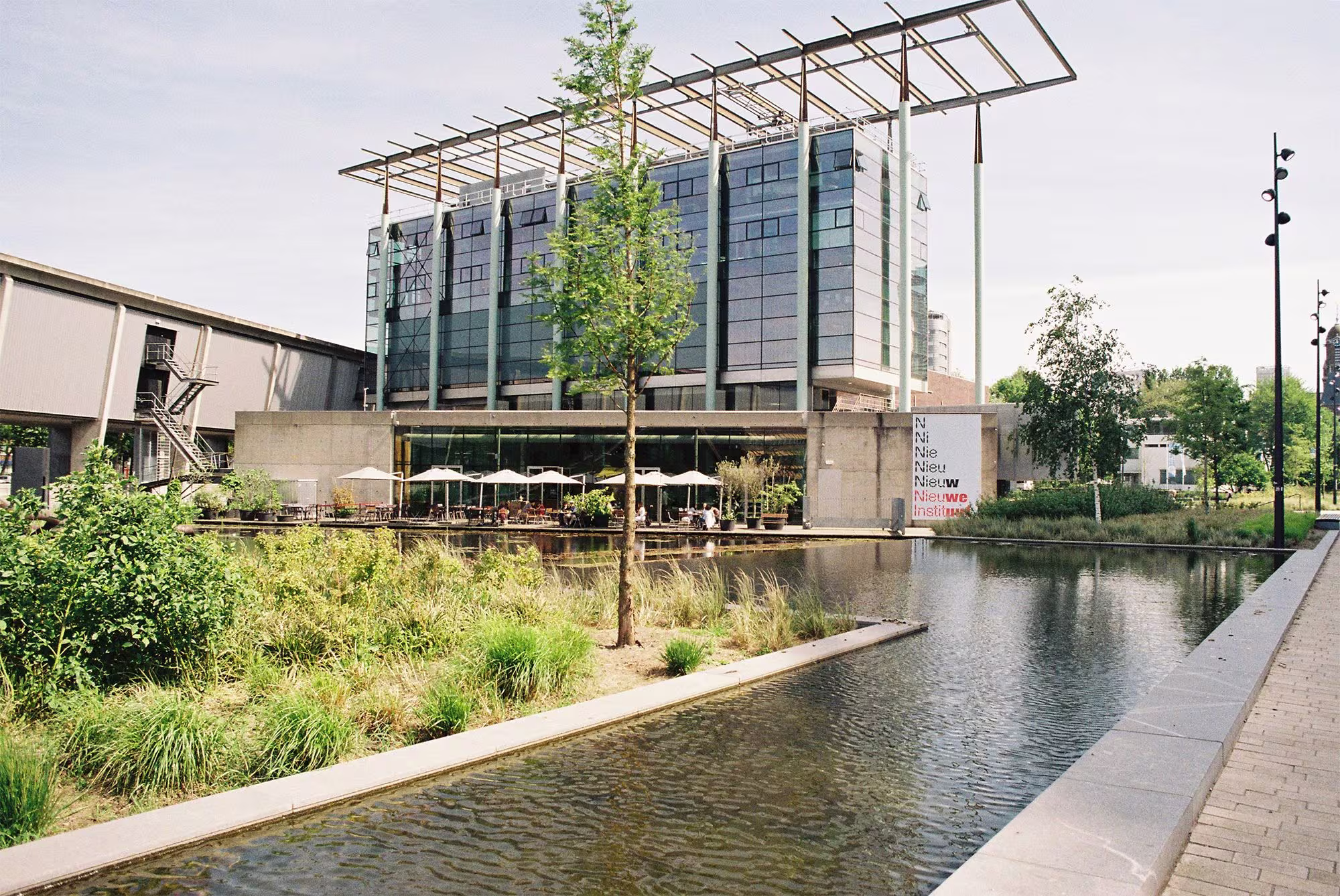Modern gemeentehuis met glazen gevel en zonnepanelen aan een waterpartij met groene beplanting en wandelpad.