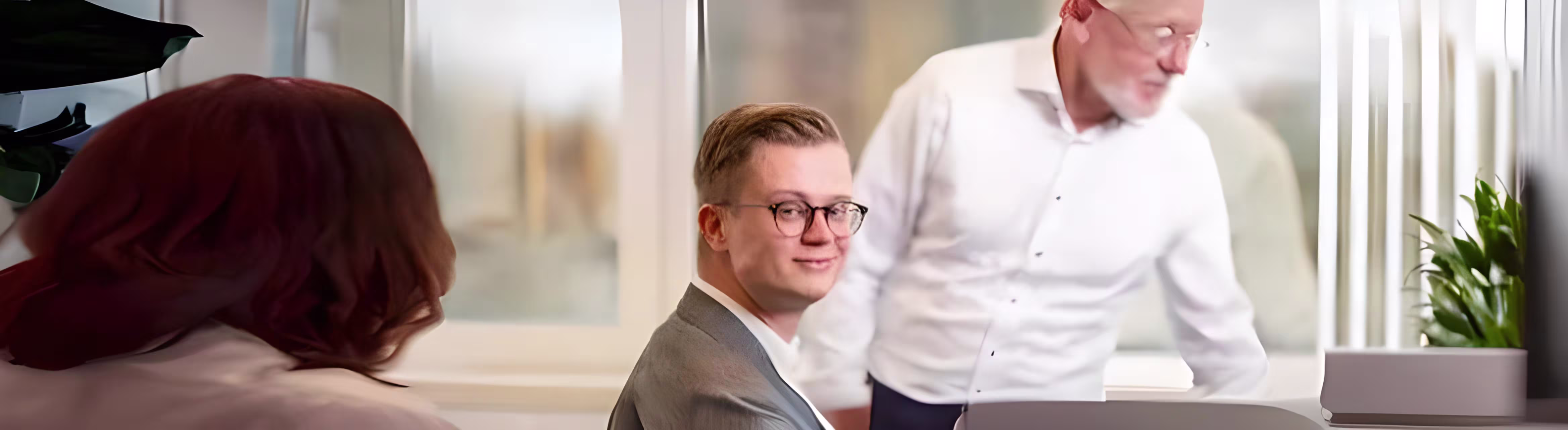 Twee mannen in pak overleggen aan een bureau in een modern kantoor, een derde persoon op de achtergrond.