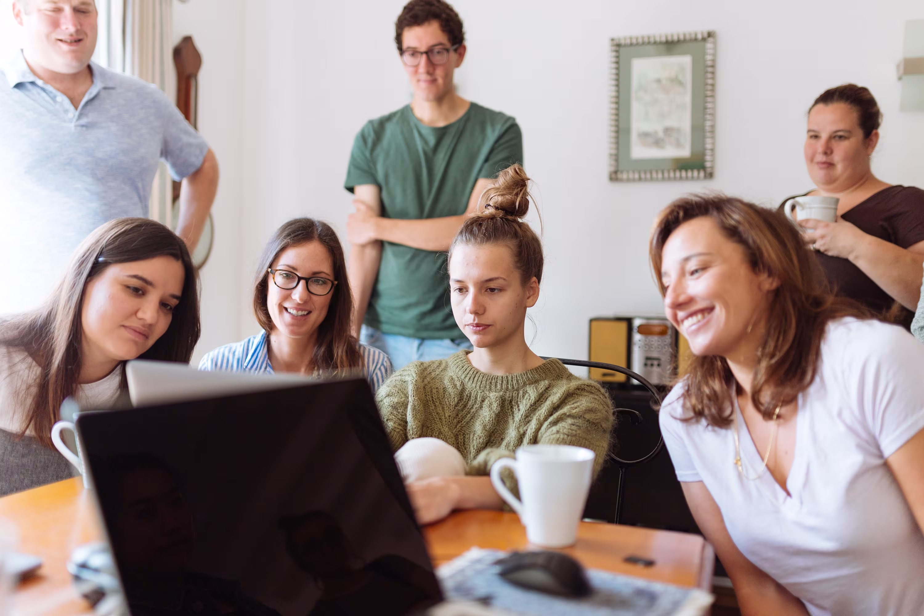 Groep jonge mensen kijkt lachend naar een laptop in een modern kantoor met koffiekopjes op tafel.