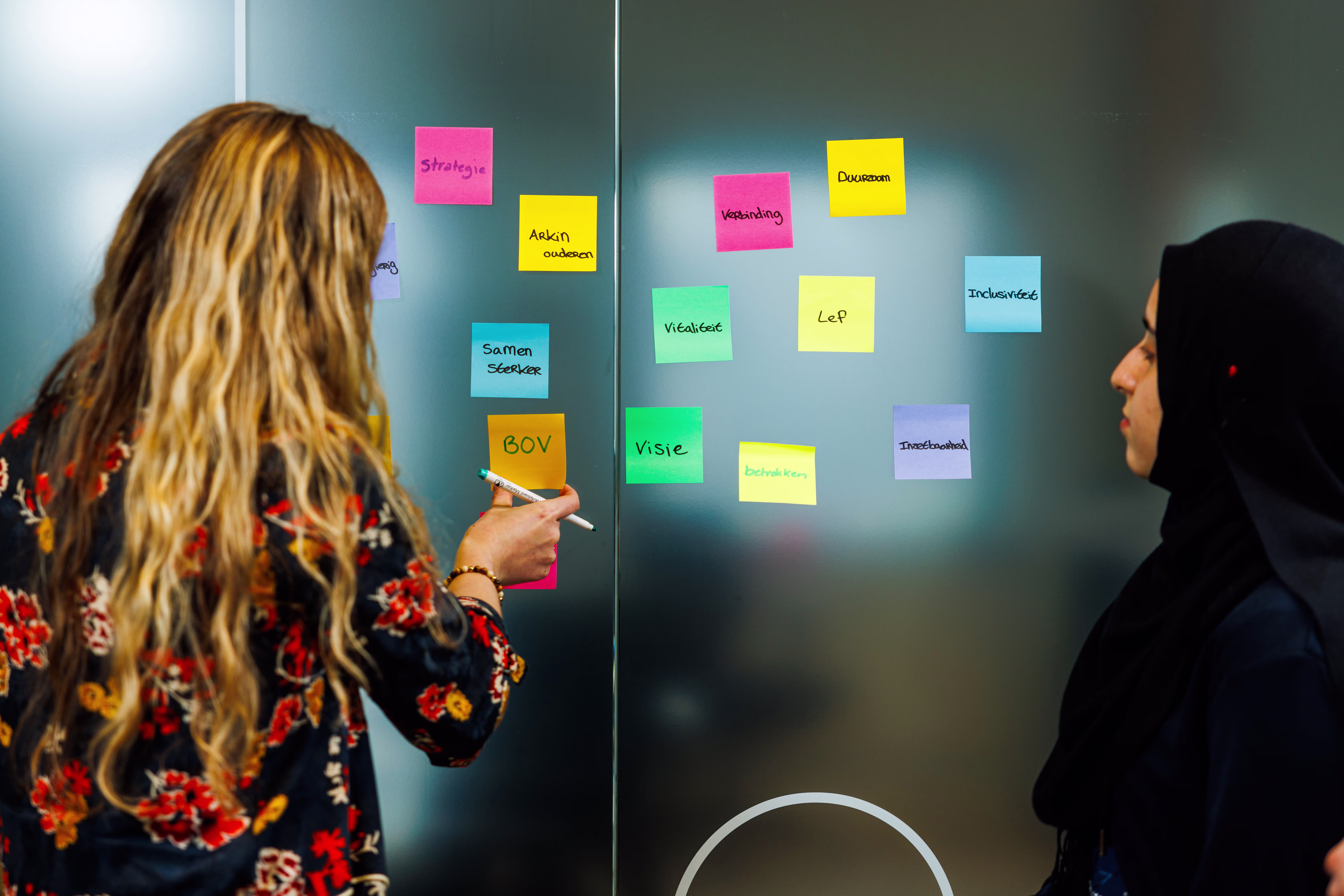 Twee vrouwelijke collega's werken samen aan een brainstormsessie met gekleurde post-it notities op een glazen wand.