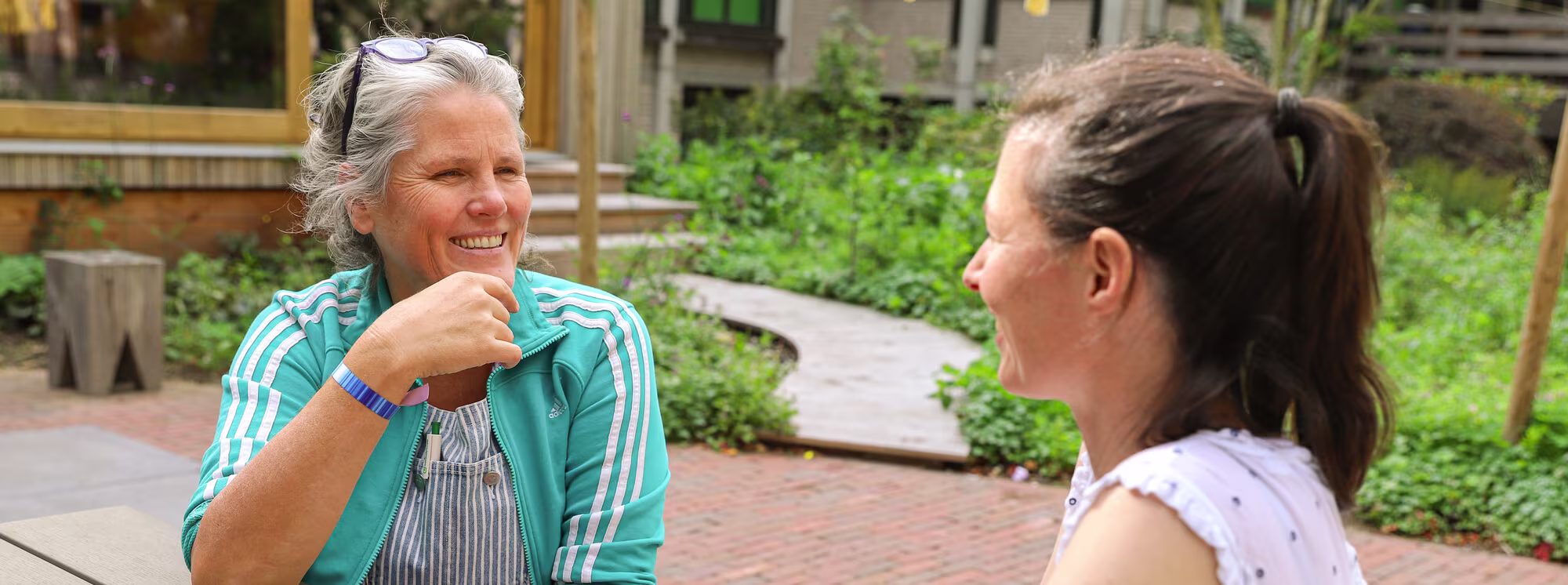 Twee vrouwen zitten buiten en hebben een gezellig gesprek in een groene tuin met een bakstenen pad.